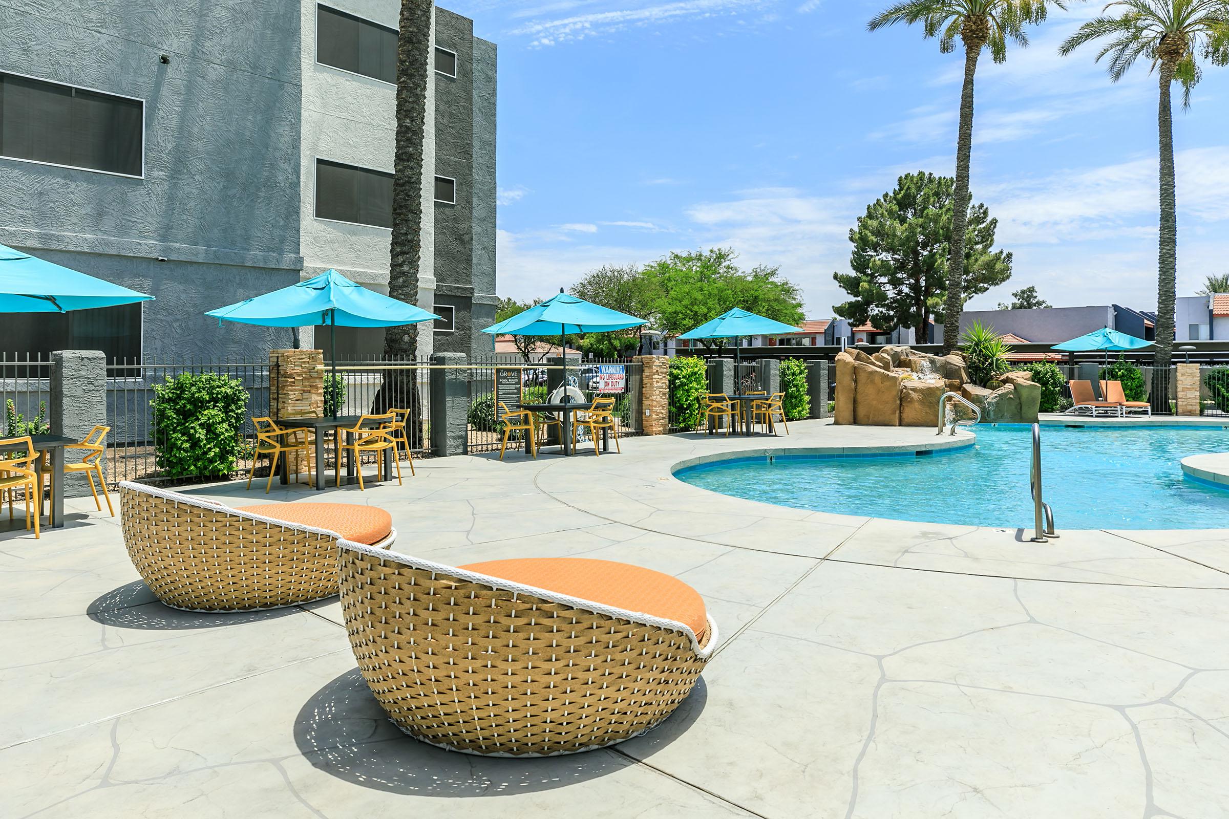 A sunny pool area featuring a blue swimming pool surrounded by lounge chairs and tables with turquoise umbrellas. There are palm trees in the background, and a rocky structure is integrated into the pool design, creating a relaxed, inviting atmosphere.