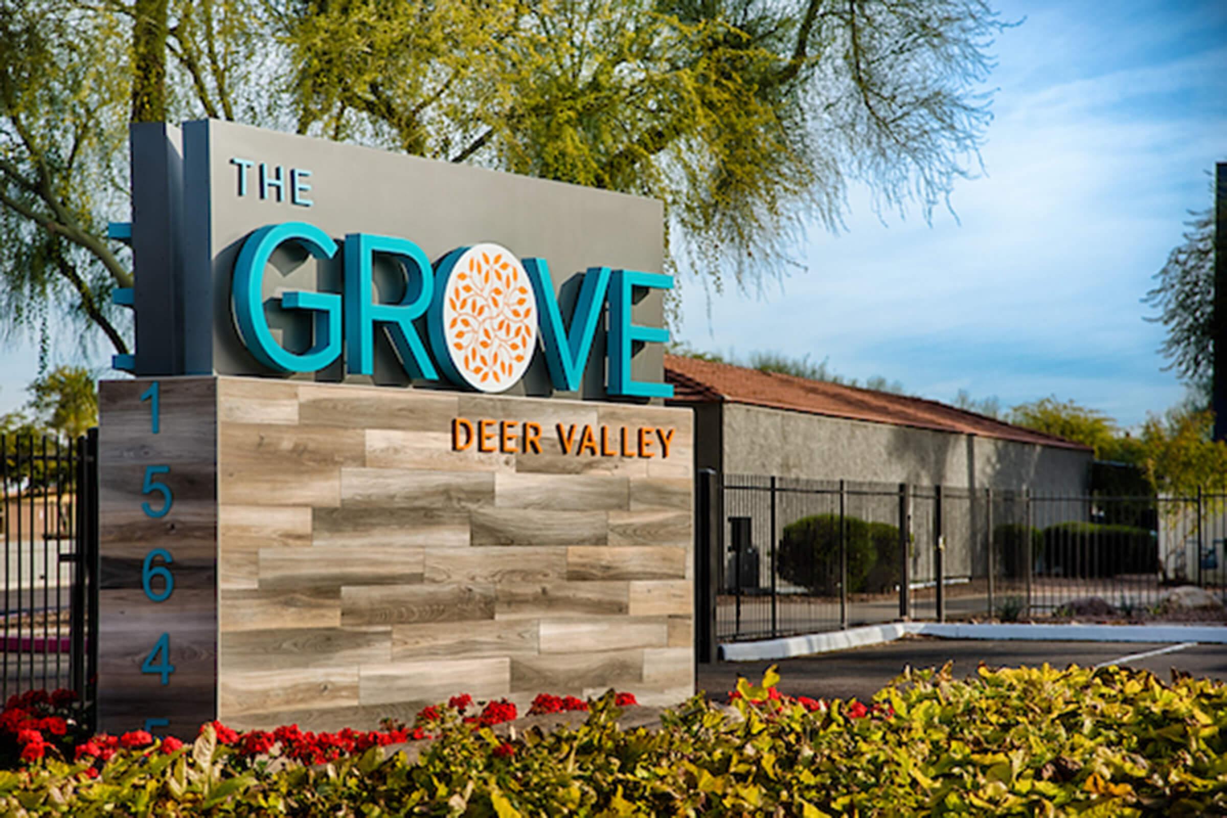 Sign displaying "The Grove" in bold blue letters, with a stylized orange tree logo, situated in Deer Valley. The sign is set against a backdrop of landscaped greenery and colorful flowers, with a building visible in the background. The address "1564" is also featured prominently.