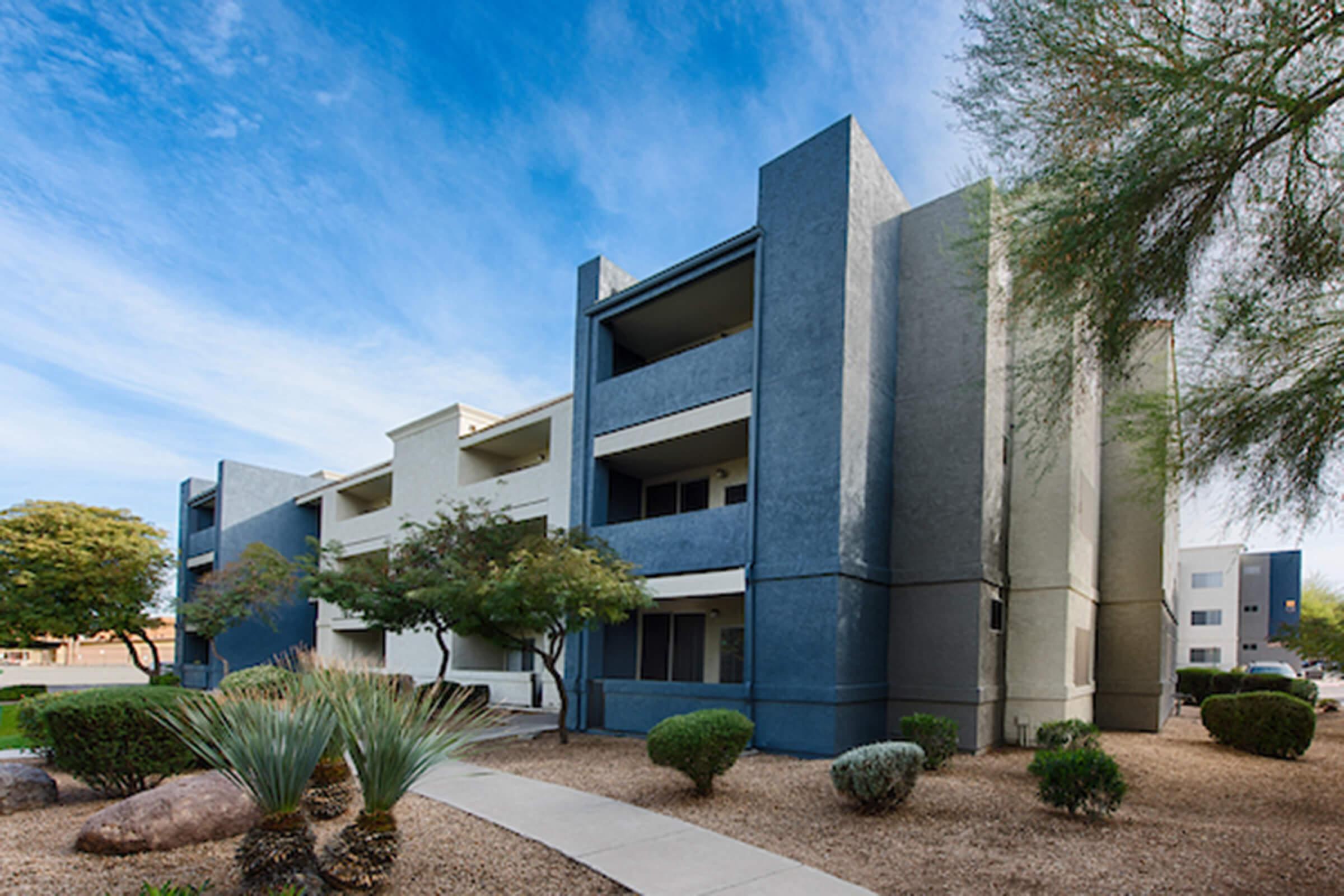 A modern apartment building with a minimalist design, featuring three levels and a combination of light and dark grey walls. Lush green landscaping, including shrubs and small trees, surrounds the building, and there’s a clear blue sky above, enhancing the serene atmosphere.