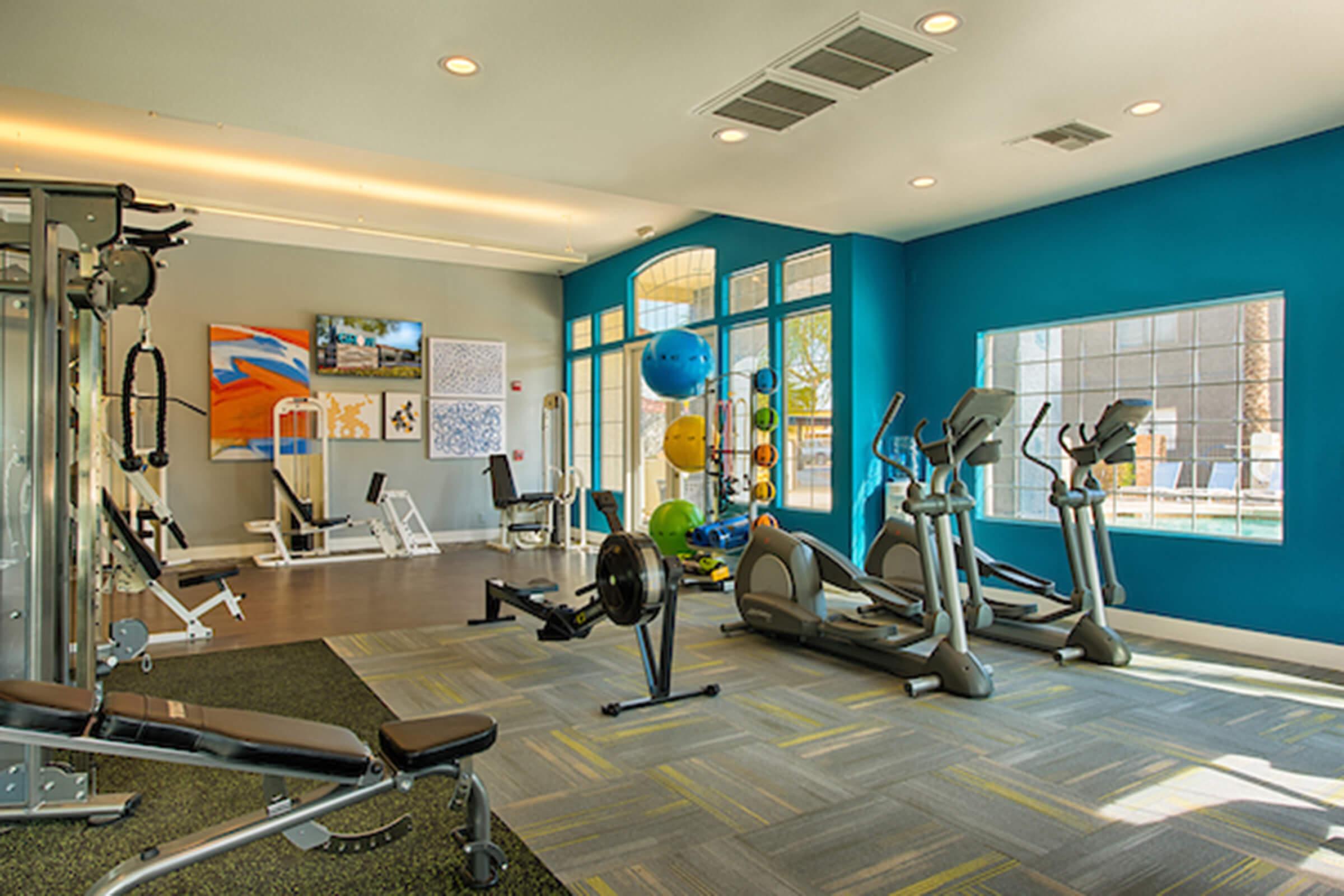 A well-lit gym featuring various exercise equipment, including treadmills, ellipticals, and weightlifting machines. The walls are painted in bright colors, and there are large windows allowing natural light to enter. Fitness accessories like exercise balls are visible, creating an inviting workout atmosphere.