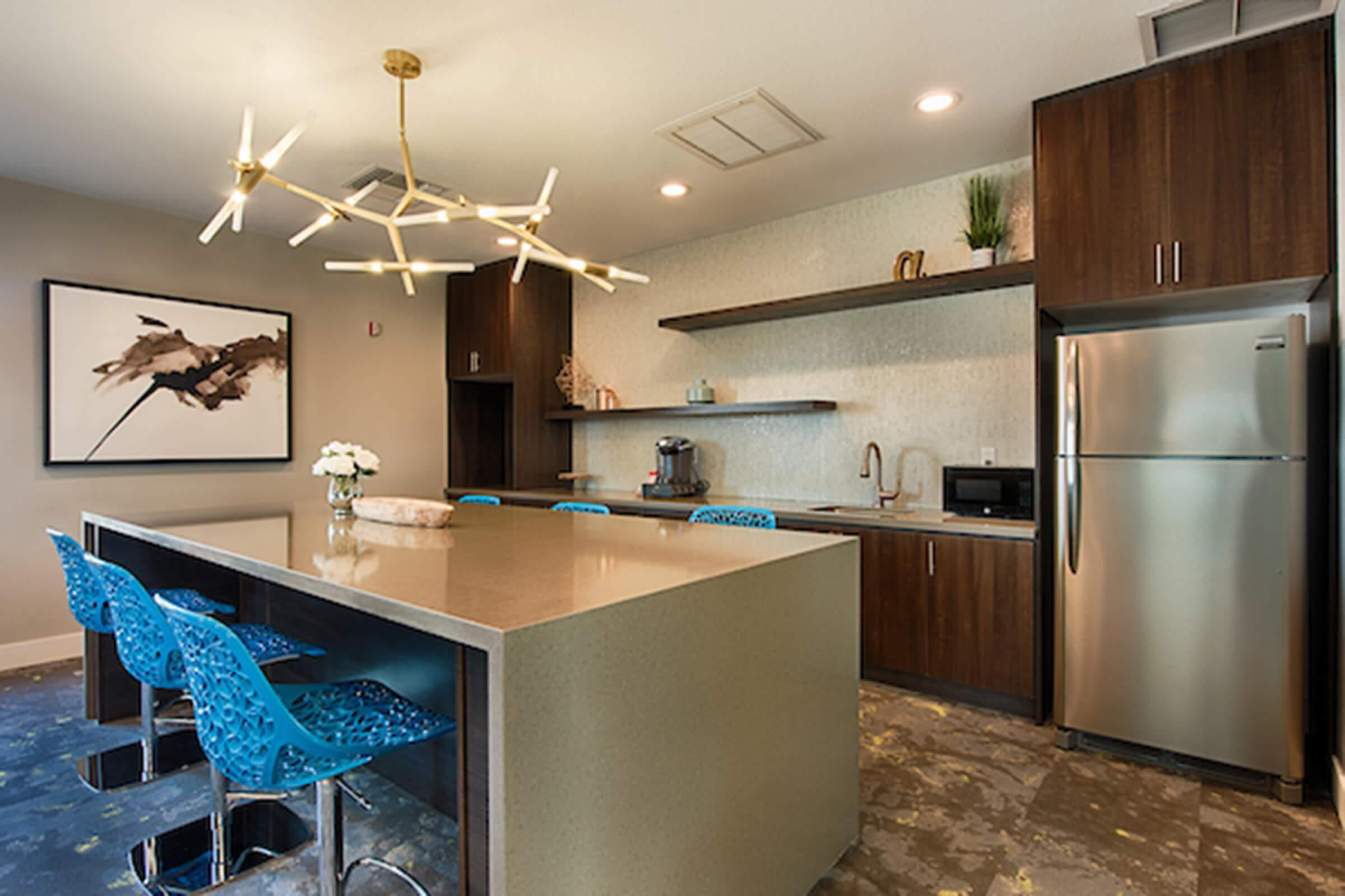 Modern kitchen interior featuring a large island with a sleek countertop, blue patterned bar stools, and contemporary lighting fixtures. The cabinetry is dark wood, complemented by open shelving and a stainless steel refrigerator. Wall decor includes an abstract painting, and the space has a clean, elegant design.