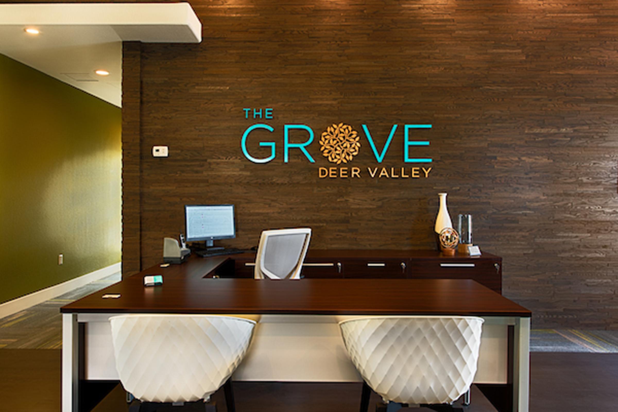 Reception area featuring a sleek desk with two modern white chairs, a computer, and decorative items. The wall behind displays the text "THE GROVE DEER VALLEY" in large, stylized letters with a floral design, set against a textured brown background.