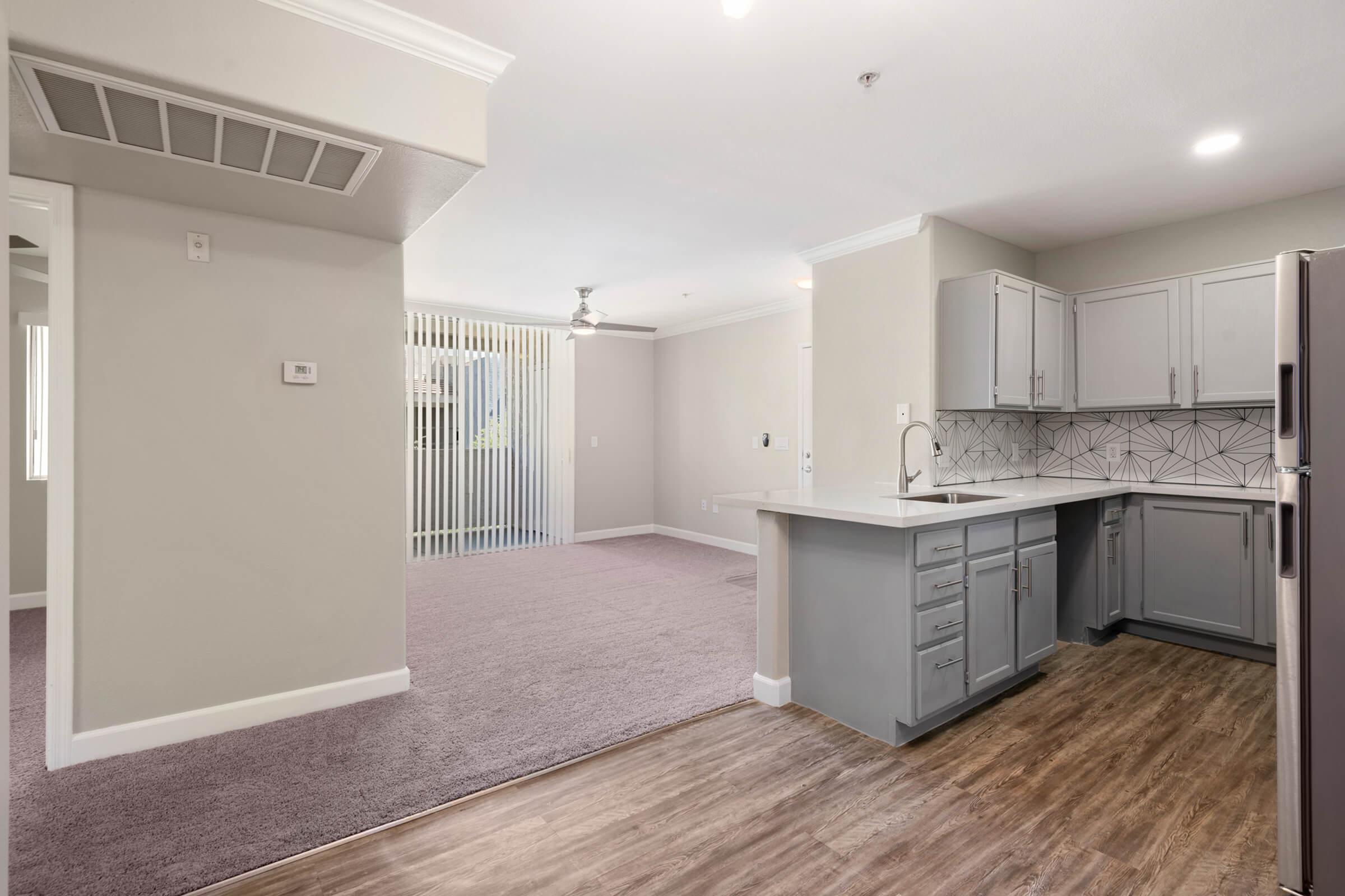 A modern apartment interior featuring an open-concept living area and kitchen. The space includes light gray walls, a carpeted living room, and a kitchen with sleek cabinets and a patterned backsplash. The area is well-lit with natural light and has a ceiling fan.