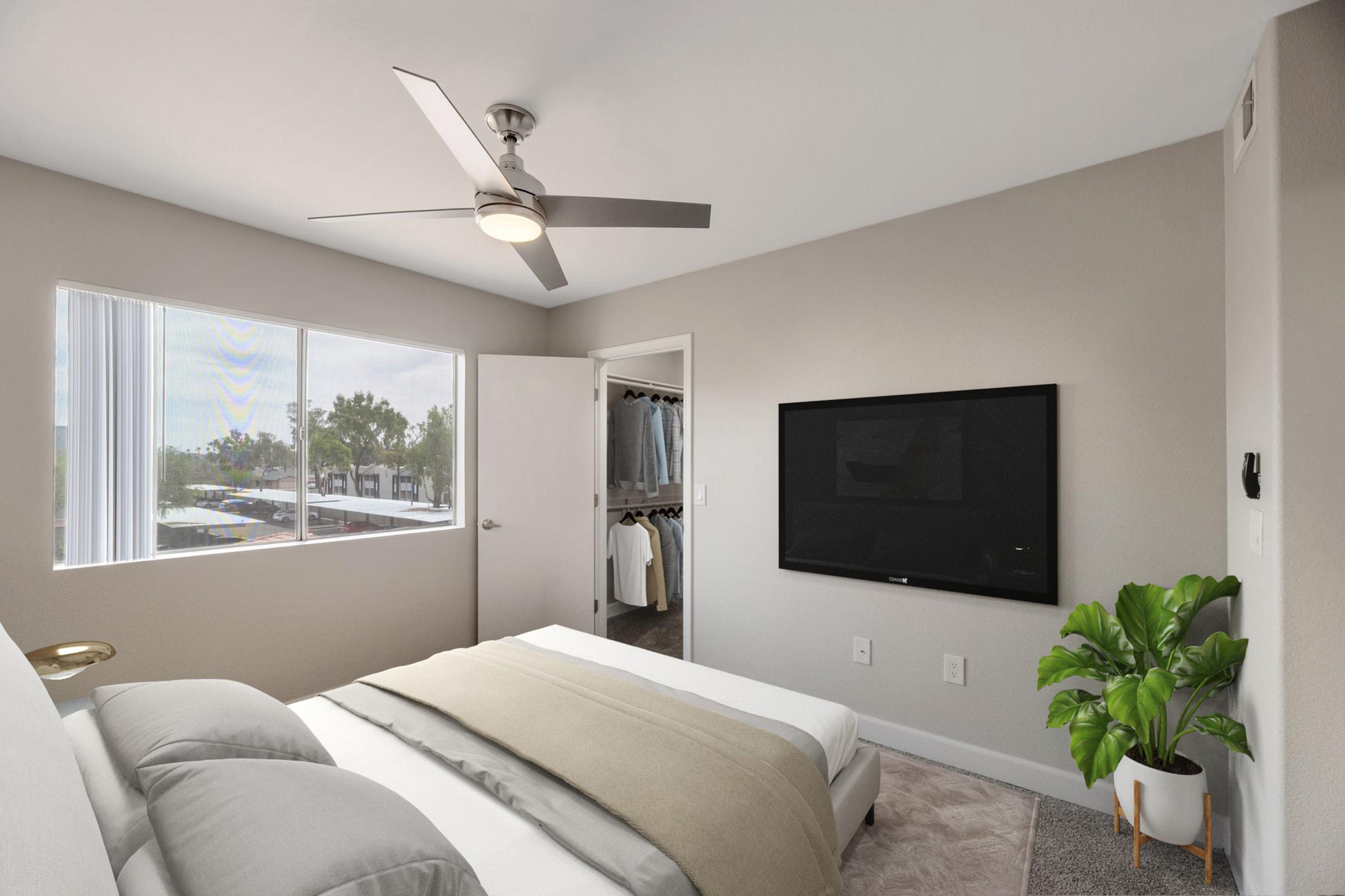 A modern bedroom featuring a queen-sized bed with neutral bedding, a ceiling fan, a large window providing natural light, and a potted plant in the corner. A flat-screen TV is mounted on the wall opposite the bed, and a closet is partially visible with clothes hanging inside. The decor is minimalistic and stylish.