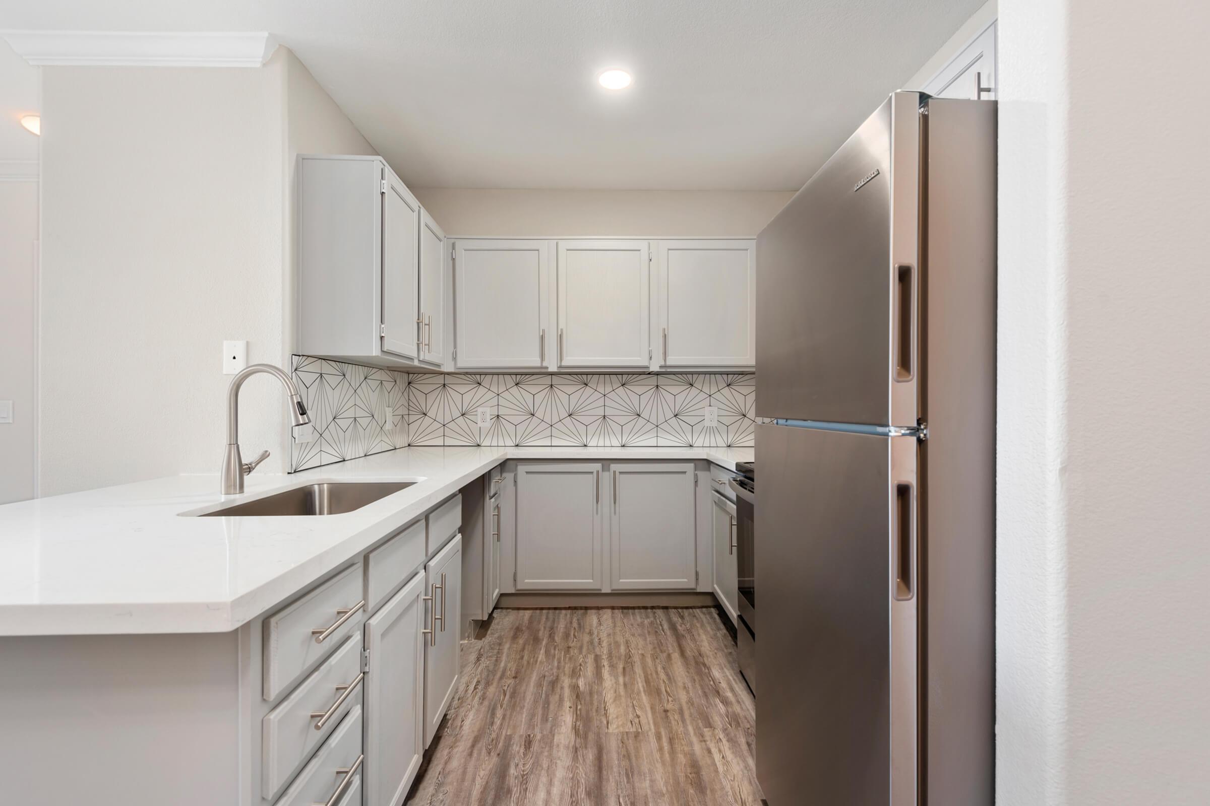 Modern kitchen featuring light gray cabinets, a stainless steel refrigerator, and a large white countertop. The backsplash showcases a geometric pattern, and the flooring is made of wood-like materials. Brightly lit with recessed lighting, the space has a clean and contemporary aesthetic.