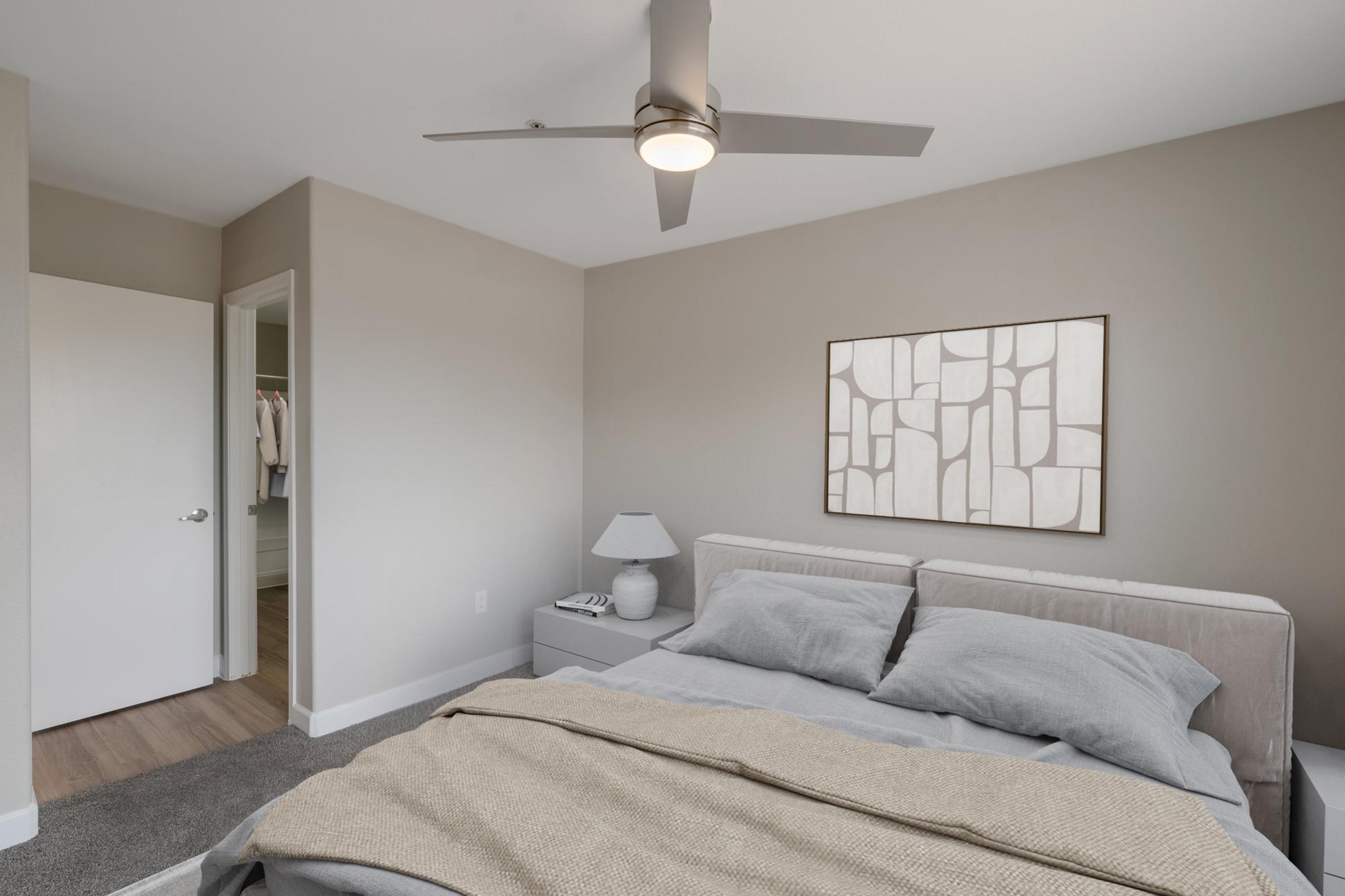 A cozy bedroom featuring a neatly made bed with gray linens, a decorative throw, and a modern lamp on a bedside table. A minimalist wall art piece hangs above the bed. The room is painted in soft neutral tones, with a ceiling fan and an open door leading to a closet space. Natural light brightens the space.