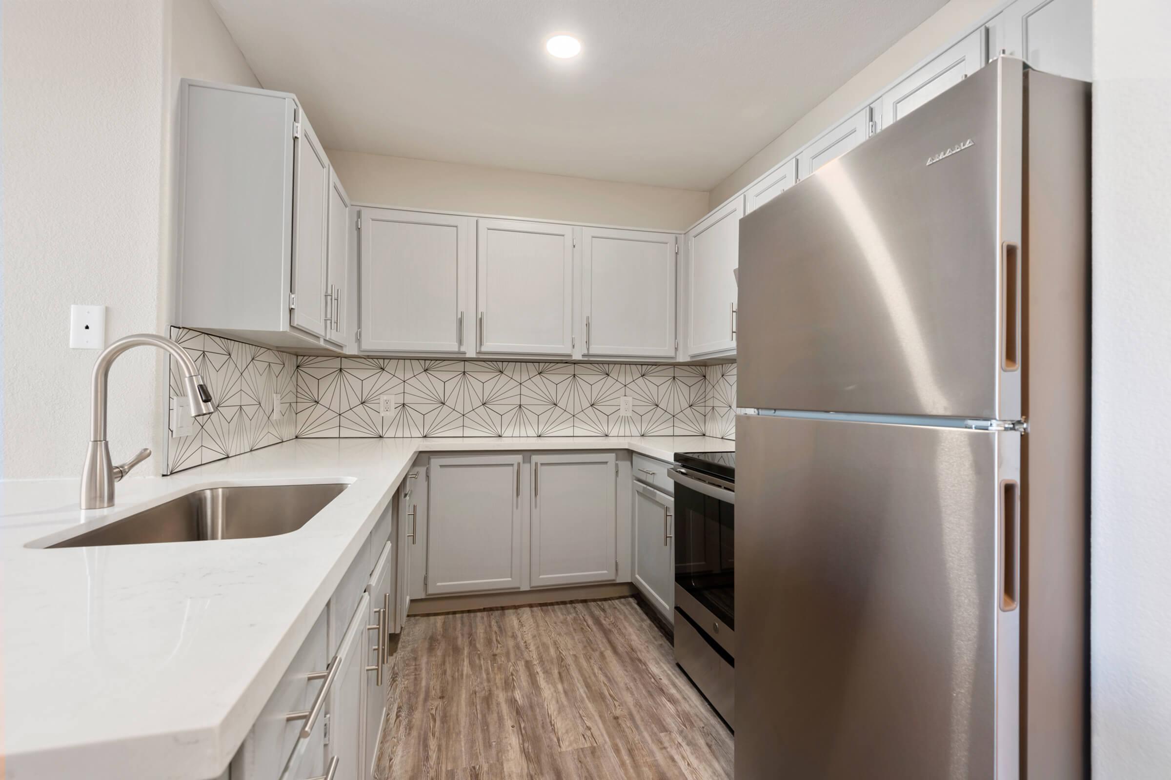 A modern kitchen featuring light gray cabinets, a stainless steel refrigerator, and a black stove. The countertops are white with a subtle texture, and the backsplash showcases a geometric design in neutral tones. The flooring is light wood, enhancing the bright and airy feel of the space.