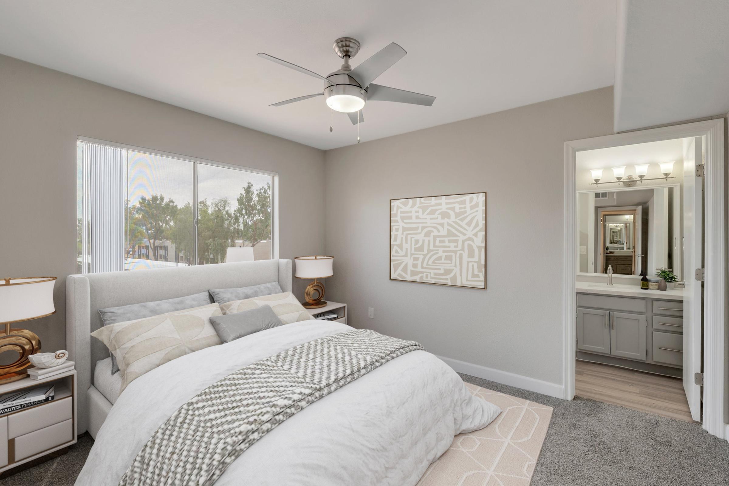 A modern bedroom featuring a light-colored bed with decorative pillows, a ceiling fan, and bedside lamps. A large window offers natural light, and there is a patterned rug on the floor. In the background, a bathroom is visible with vanity lighting and a mirror.