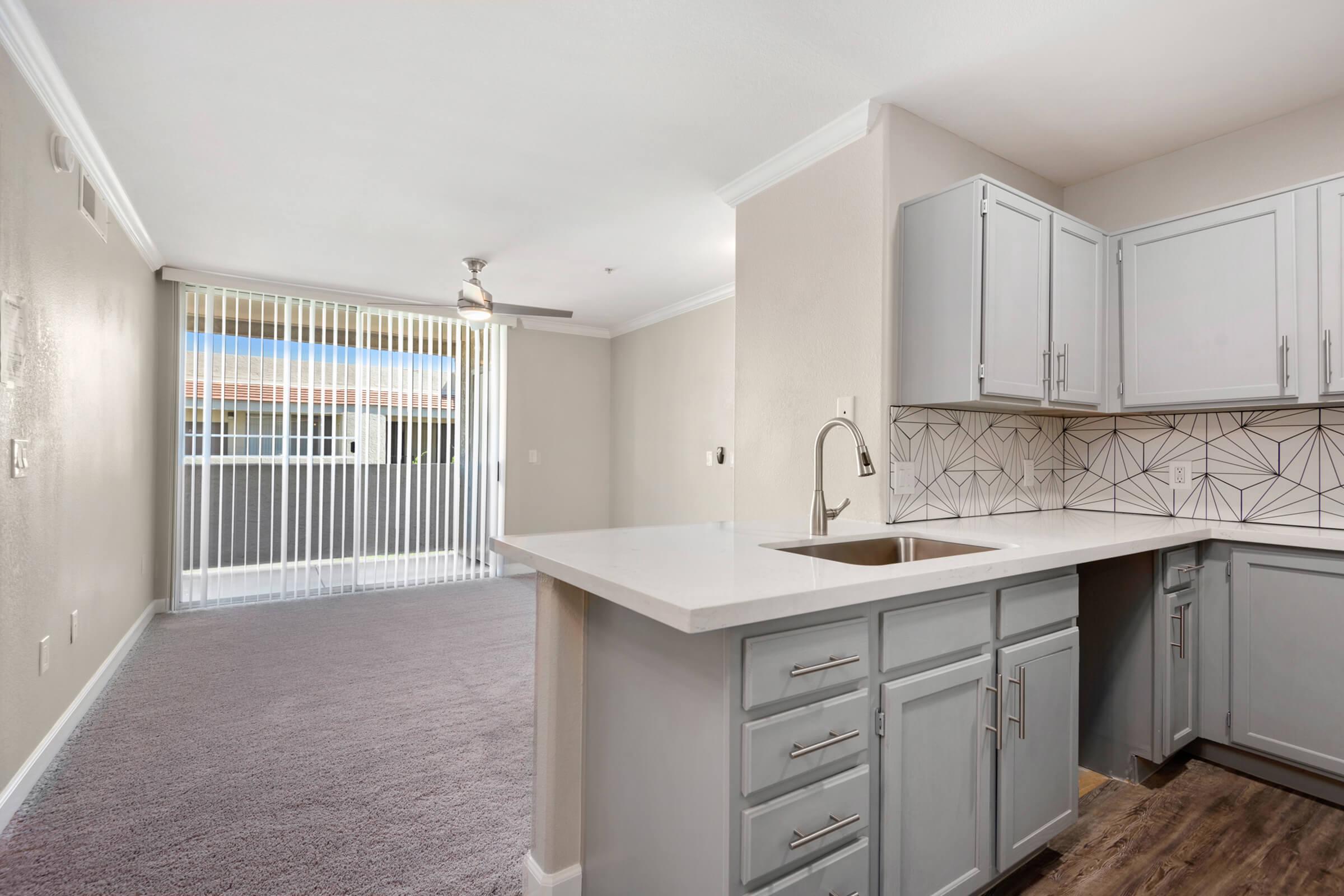 Modern kitchen with light gray cabinets and a white countertop, featuring a sink and geometric backsplash. The open layout transitions to a living area with large windows covered by vertical blinds, revealing a view outside. The floor is carpeted in a neutral tone, creating a bright and airy atmosphere.