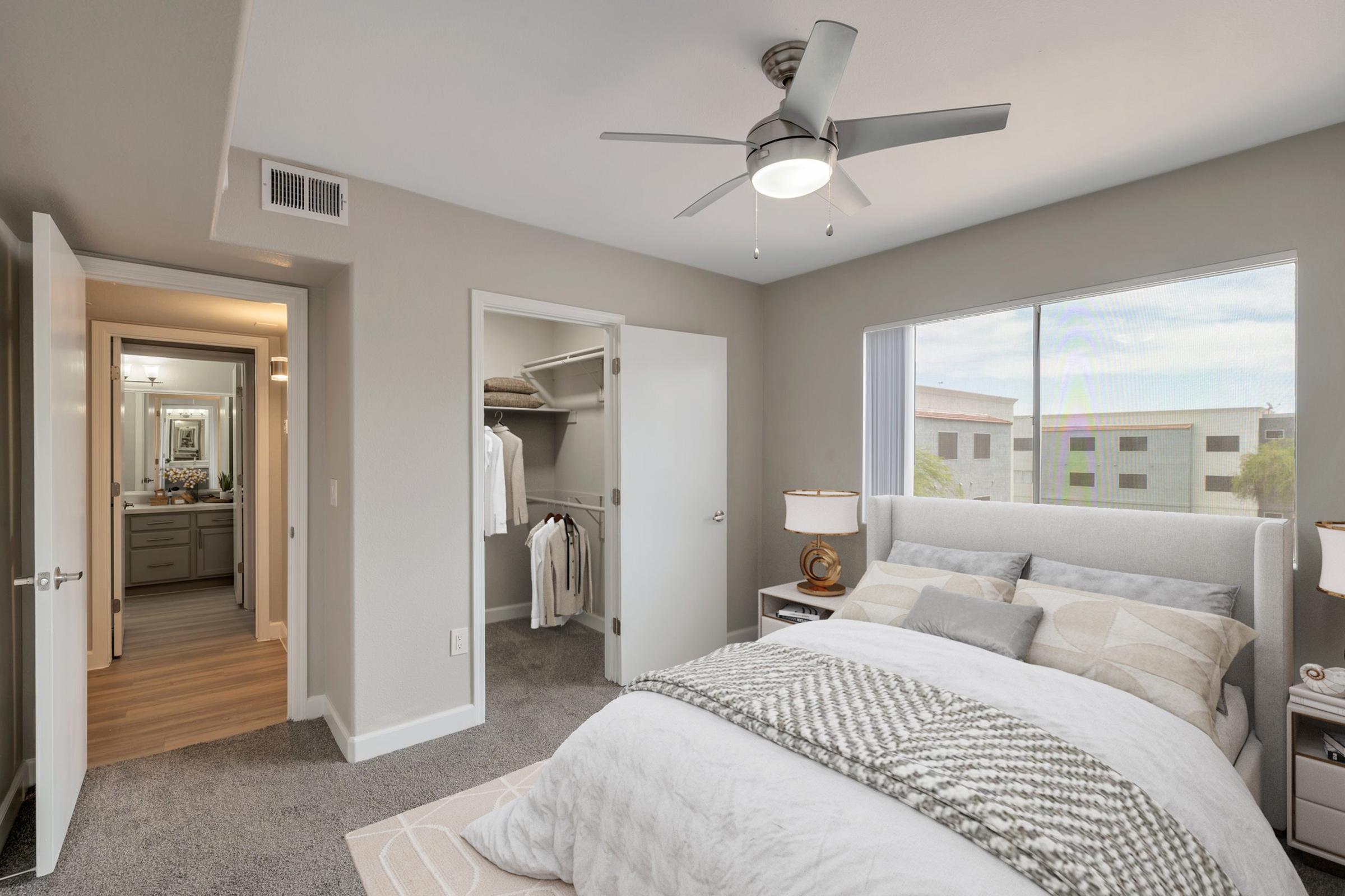 A modern bedroom featuring a cozy bed with a light-colored blanket and decorative pillows. There are two bedside lamps on nightstands, a ceiling fan, and a window allowing natural light. A doorway leads to a bathroom with a mirror, while a closet is visible, showcasing organized clothing.