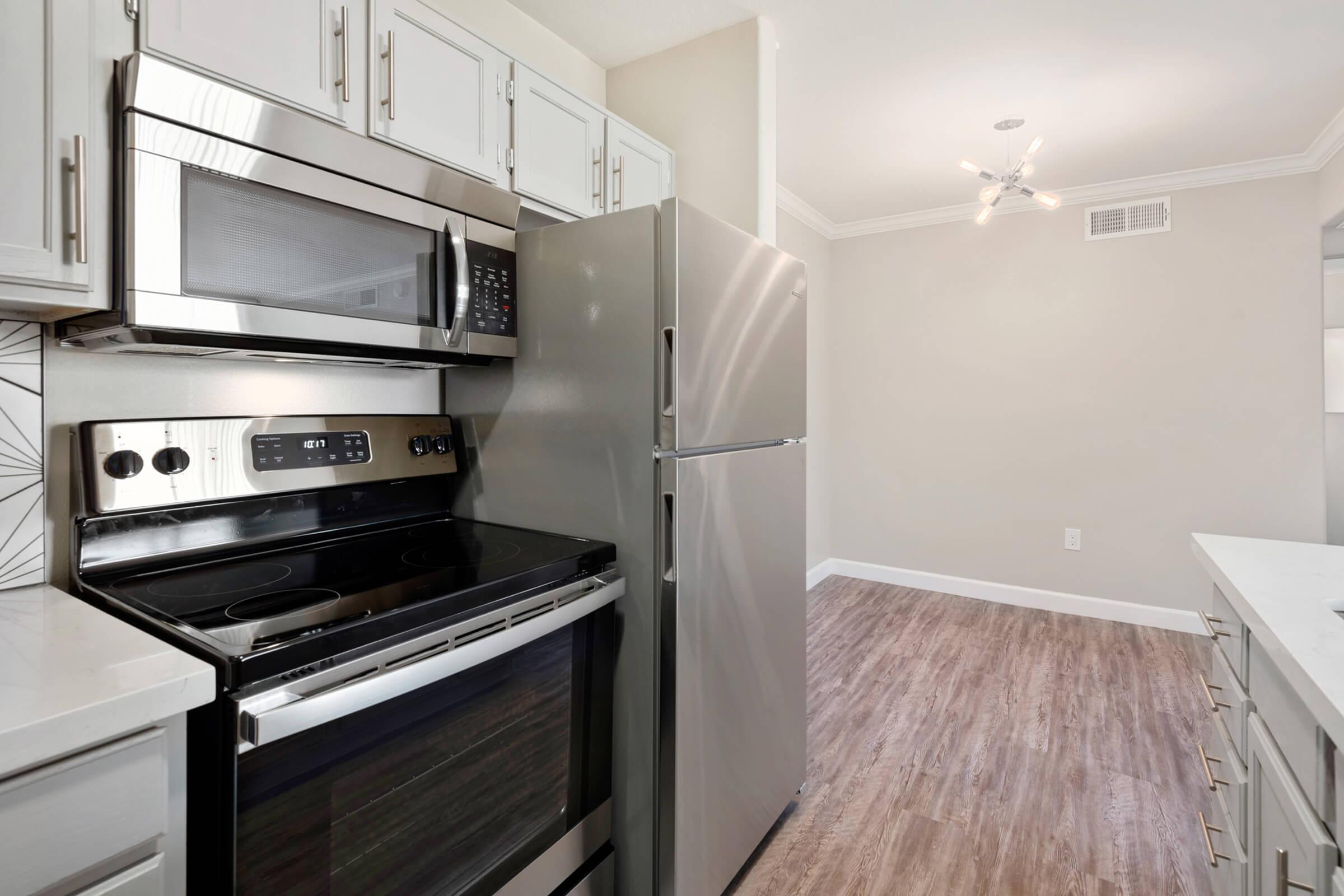 Modern kitchen featuring stainless steel appliances including an oven, microwave, and refrigerator. The cabinetry is white with sleek handles, and the floor is a light wood laminate. Soft lighting from a contemporary ceiling fixture enhances the bright, inviting atmosphere.