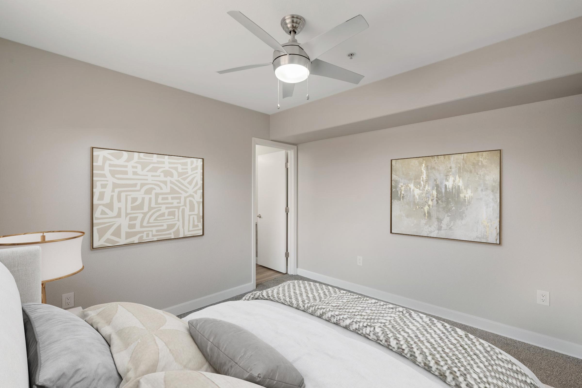 Cozy bedroom featuring a ceiling fan, a neatly made bed with decorative pillows, and two abstract artworks on light-colored walls. A door leads to another room, and soft lighting enhances the peaceful atmosphere of the space.