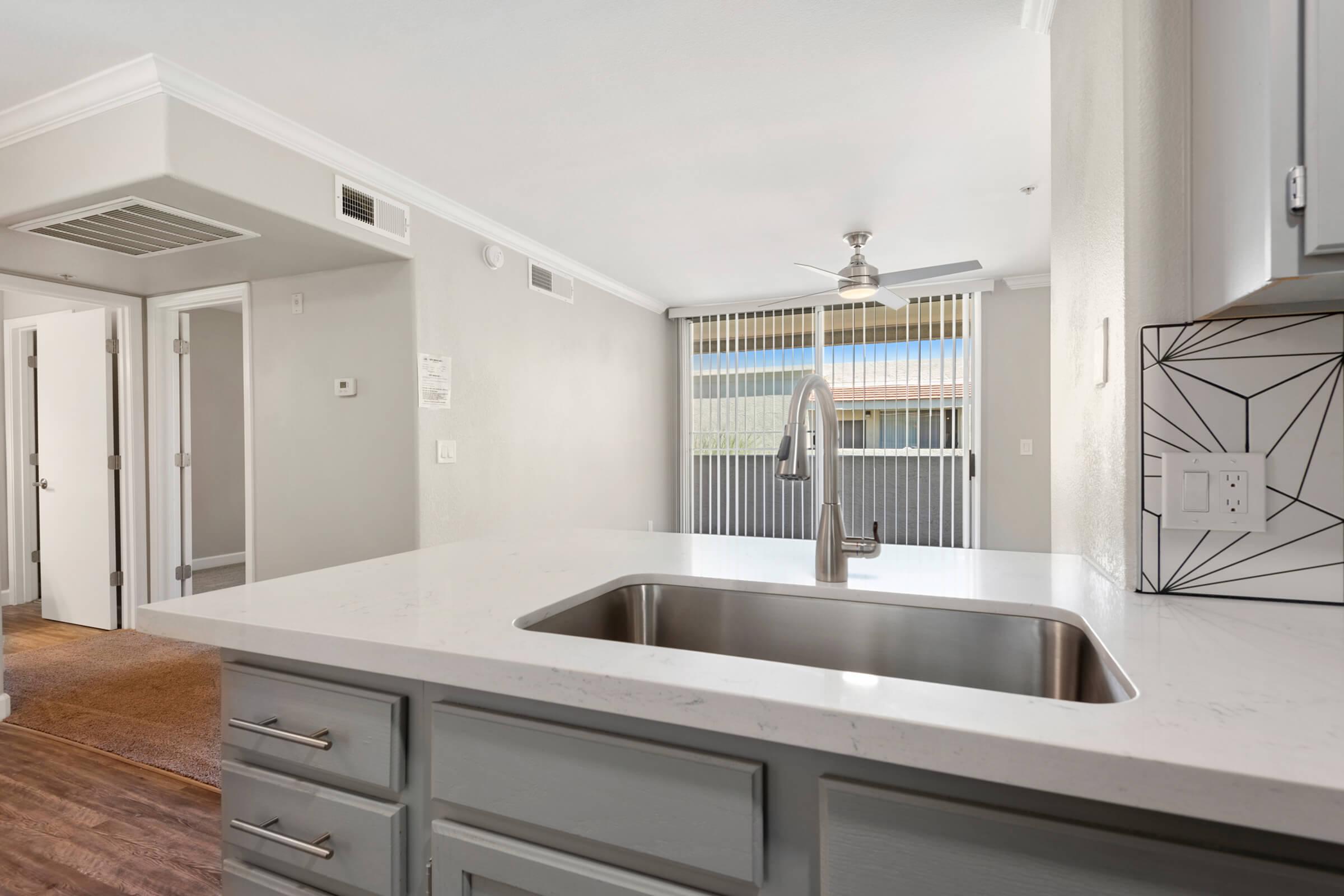 Modern kitchen with a quartz countertop and stainless steel sink, featuring gray cabinetry. In the background, a bright room with large windows and ceiling fan, and an open layout leading to another room. Soft natural light filters through sheer window coverings.