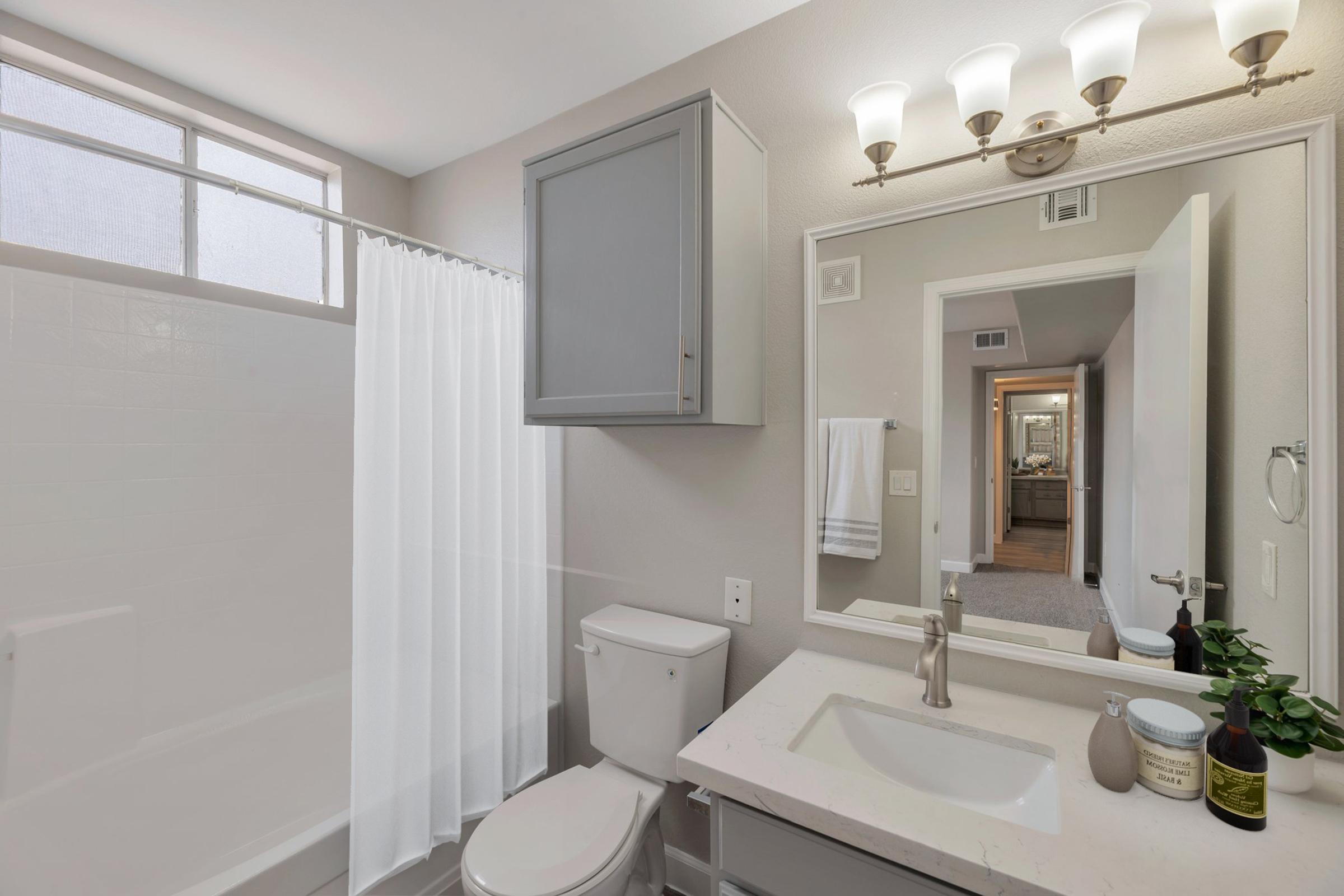 A clean, modern bathroom featuring a white bathtub with a shower curtain, a toilet, and a stylish sink with a countertop. There is a wall-mounted mirror above the sink, a bathroom cabinet, and soft lighting from four fixtures above the mirror. The design is minimalistic, with neutral tones and natural light coming through a window.