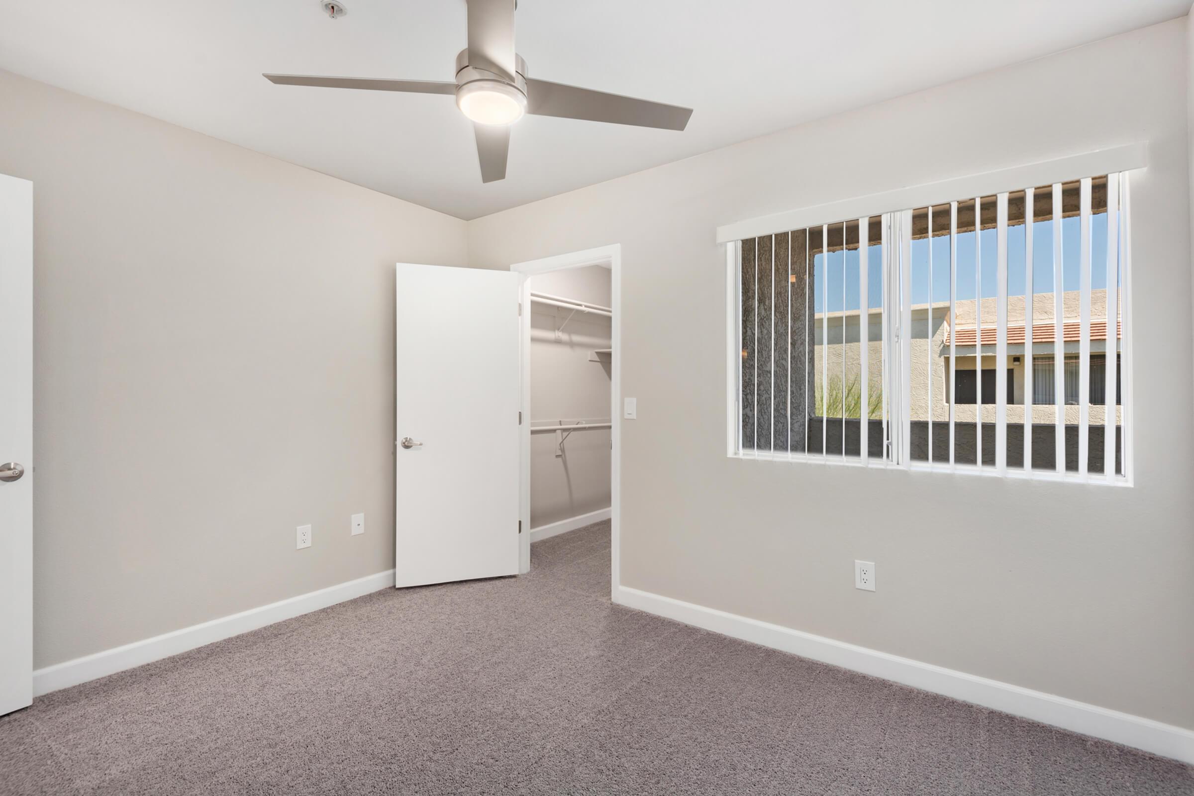 A spacious, well-lit room featuring light gray walls, a ceiling fan, and a large window with white blinds. The room includes an open door leading to a closet space with shelving. The flooring is carpeted in a soft gray, enhancing the modern aesthetic. Natural light fills the room from the window.