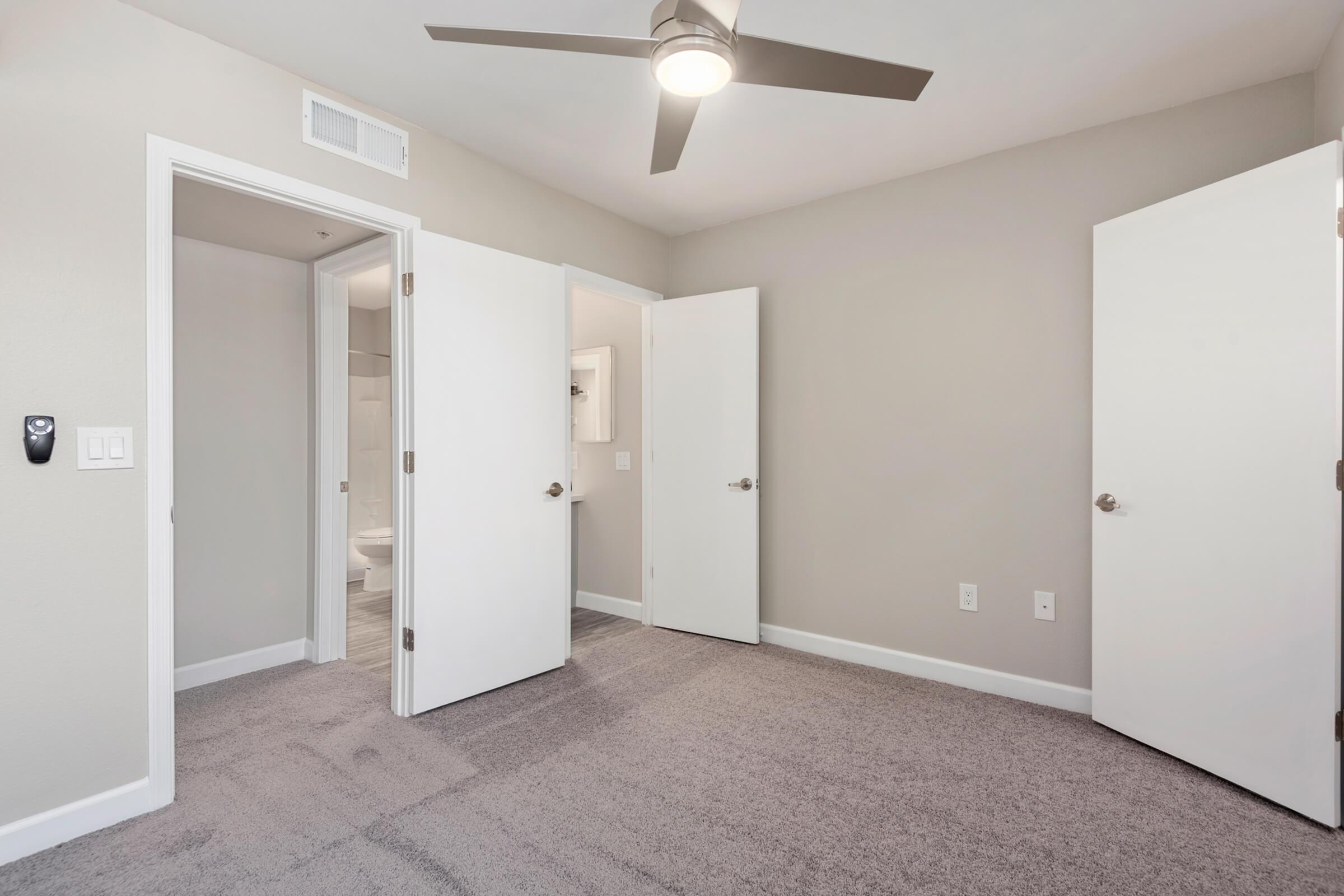 A modern, empty room with light gray walls and carpet. Two white doors are open, leading to different areas, while a ceiling fan is installed in the center. The space is well-lit and features minimalistic design elements, creating a clean and airy atmosphere. 