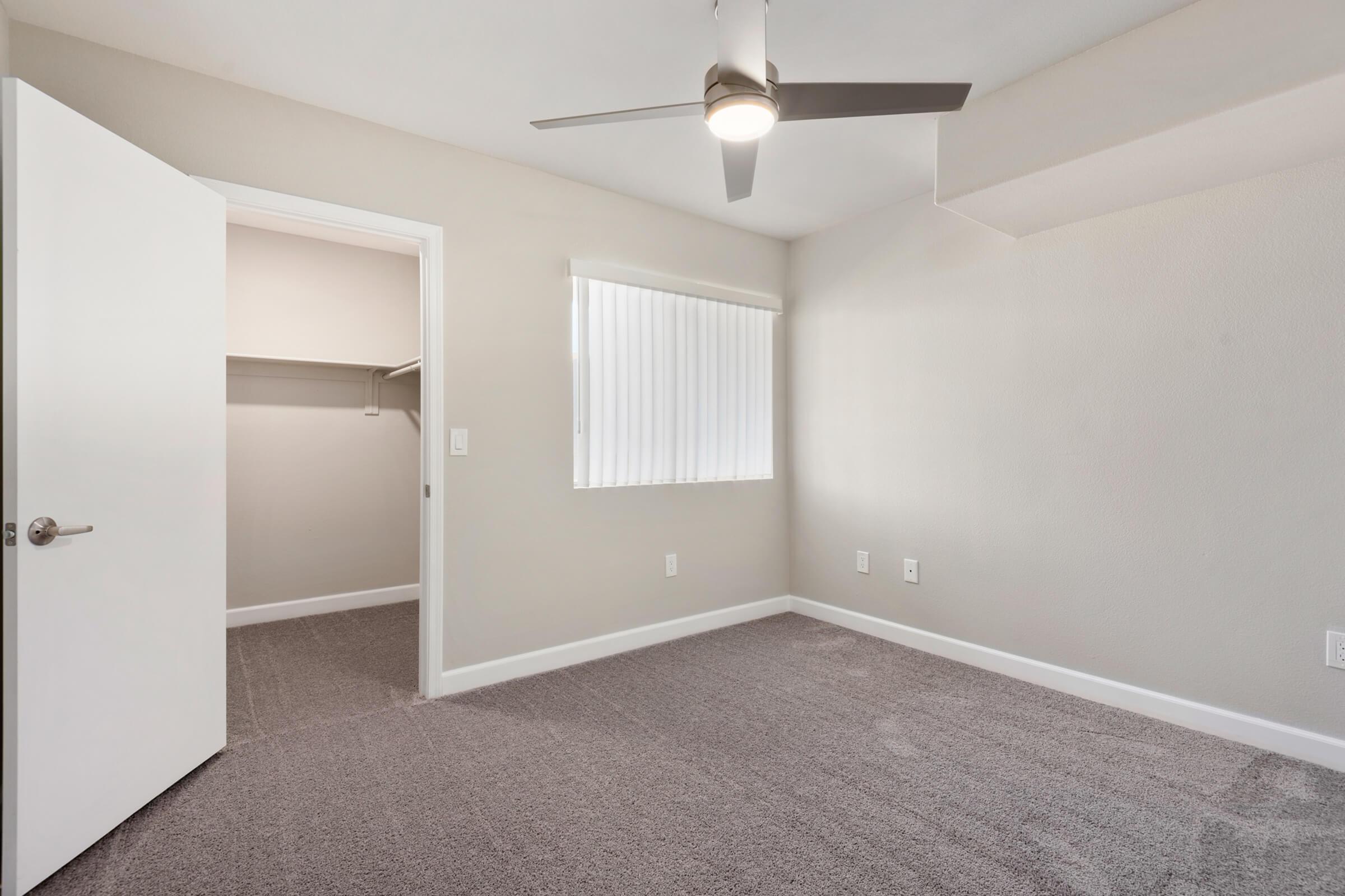 A vacant room with light gray walls and carpet. It features a ceiling fan, a window with vertical blinds, and an open door leading to a closet. The room is well-lit, creating a spacious and airy feel.