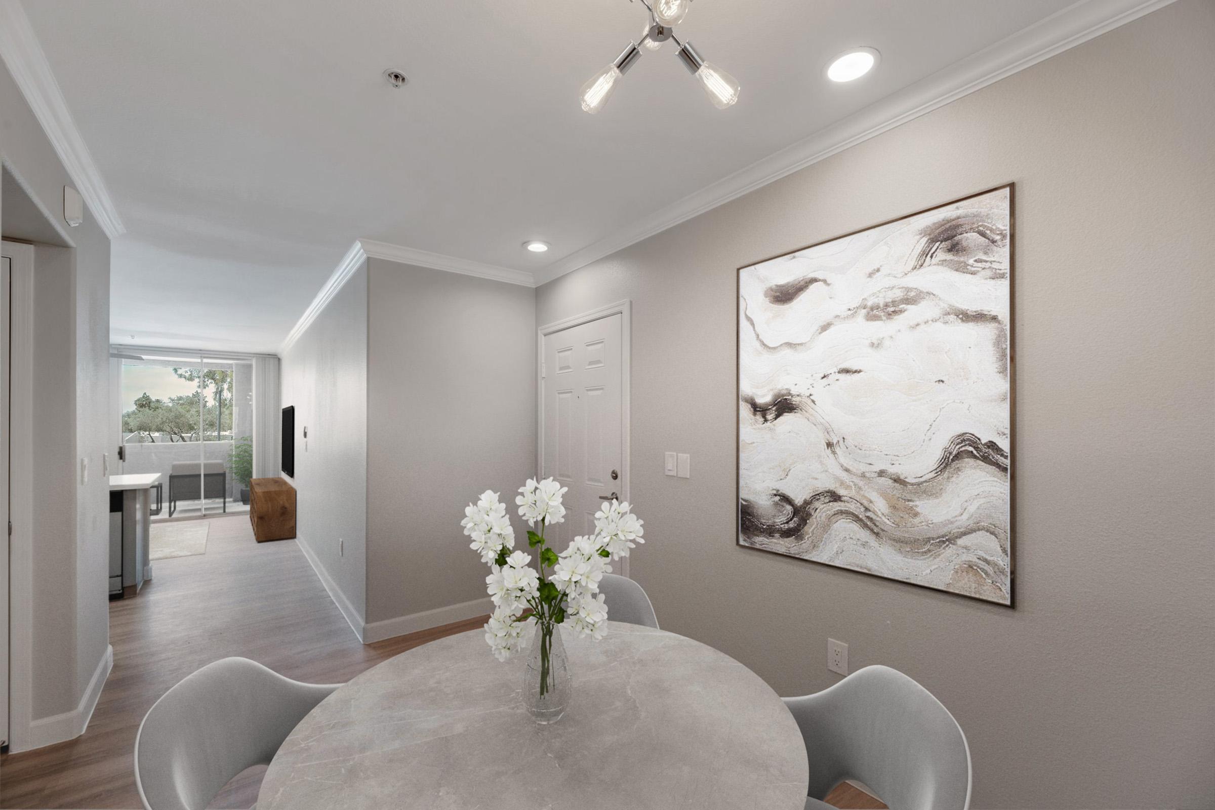 Modern dining area with a round table and vase of white flowers. A large abstract painting in neutral tones hangs on the light gray wall. The space has soft lighting from a ceiling fixture, and a doorway leads to a bright area with large windows. Light wood flooring adds warmth to the room.