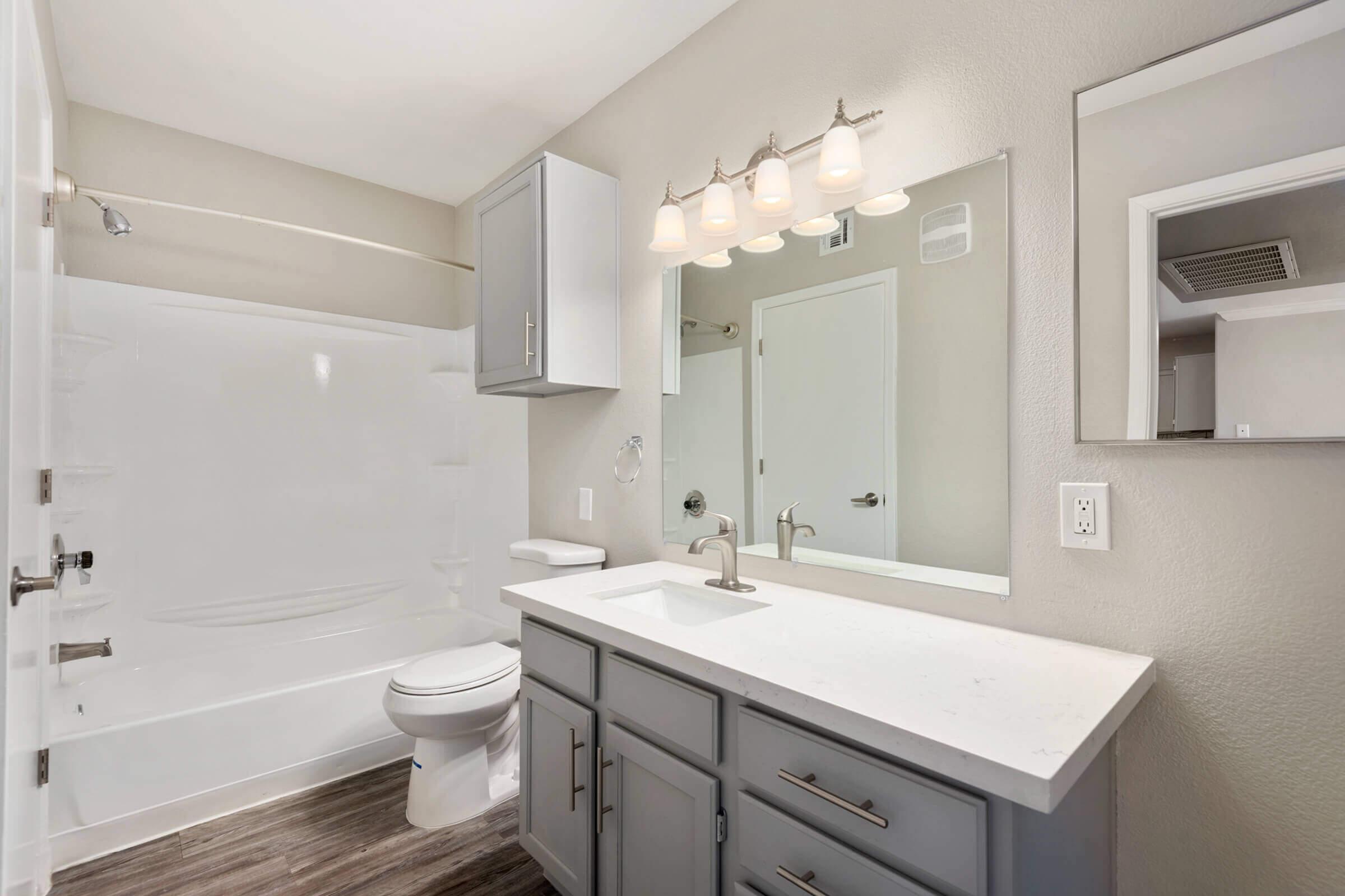 A modern bathroom featuring a white bathtub, a toilet, and a sleek gray vanity with a white countertop. Above the vanity, there are three light fixtures. The walls are painted in a light shade, and there is a large mirror above the sink. The floor has a wood-like appearance.