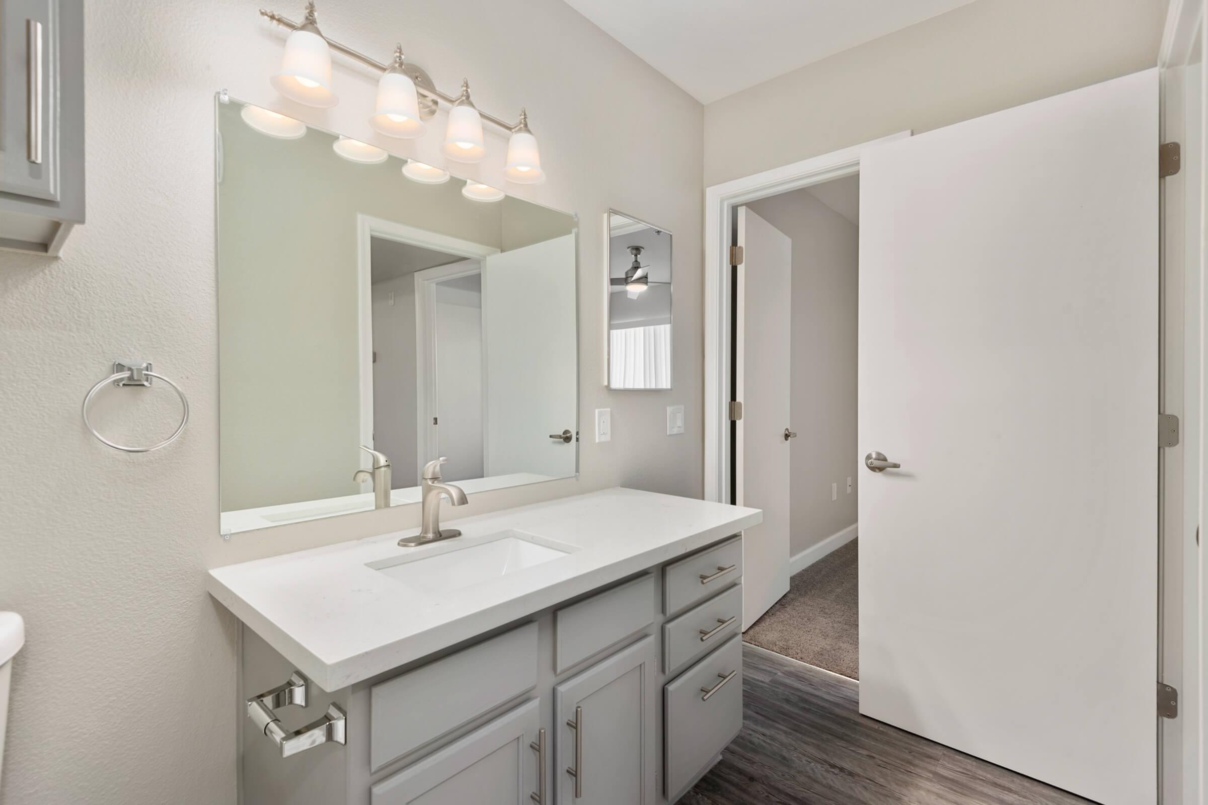 Modern bathroom featuring a light gray vanity with a white countertop and a dual mirror setup. The space is illuminated by four pendant lights above the mirrors. A doorway leads to an adjoining room, and the flooring is dark wood laminate. The walls are painted in a soft neutral tone.