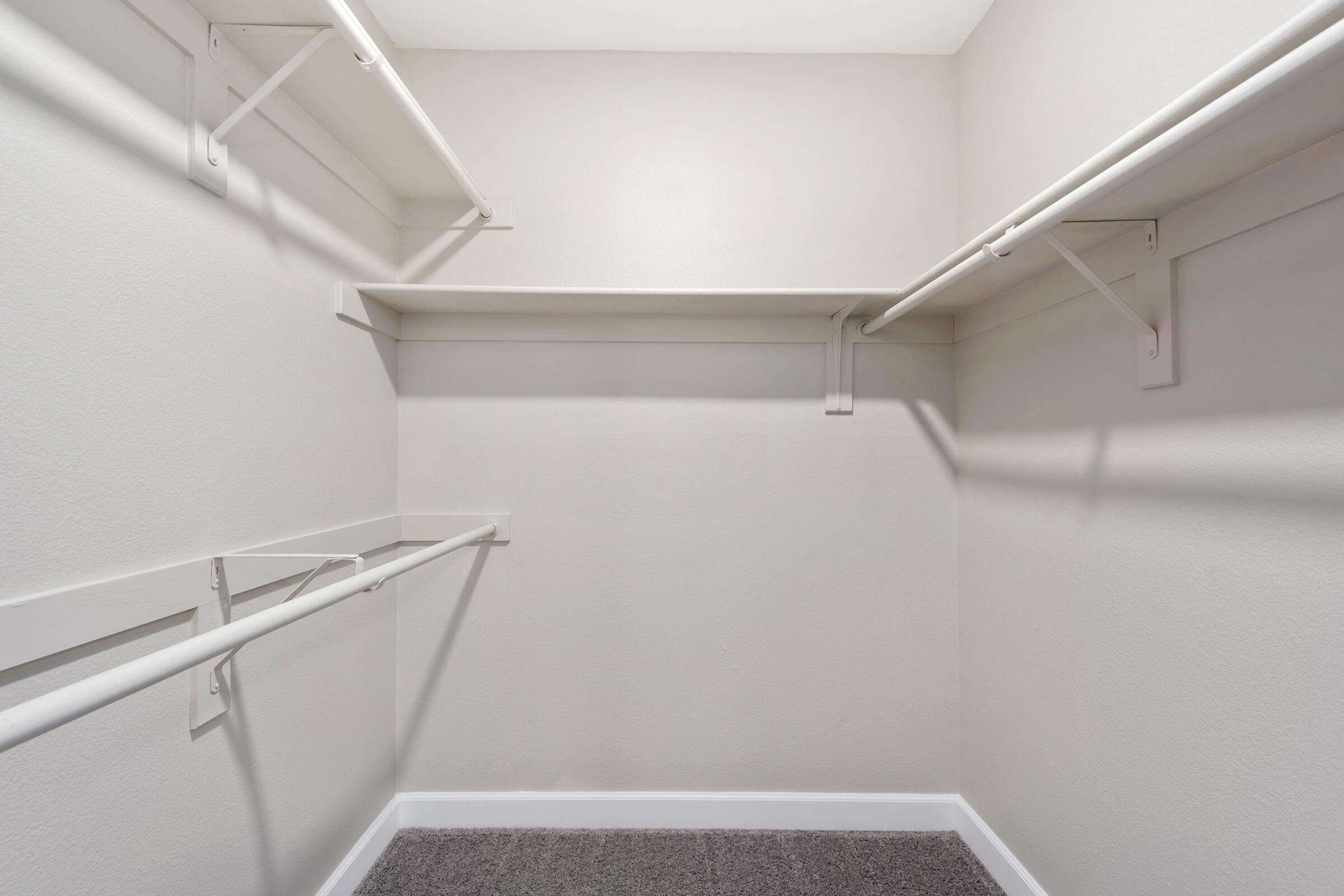 Empty closet with light gray walls and a carpeted floor. There are several white shelves and hanging rods along the walls, providing storage space, but no items are visible. The overall atmosphere is clean and minimalistic.