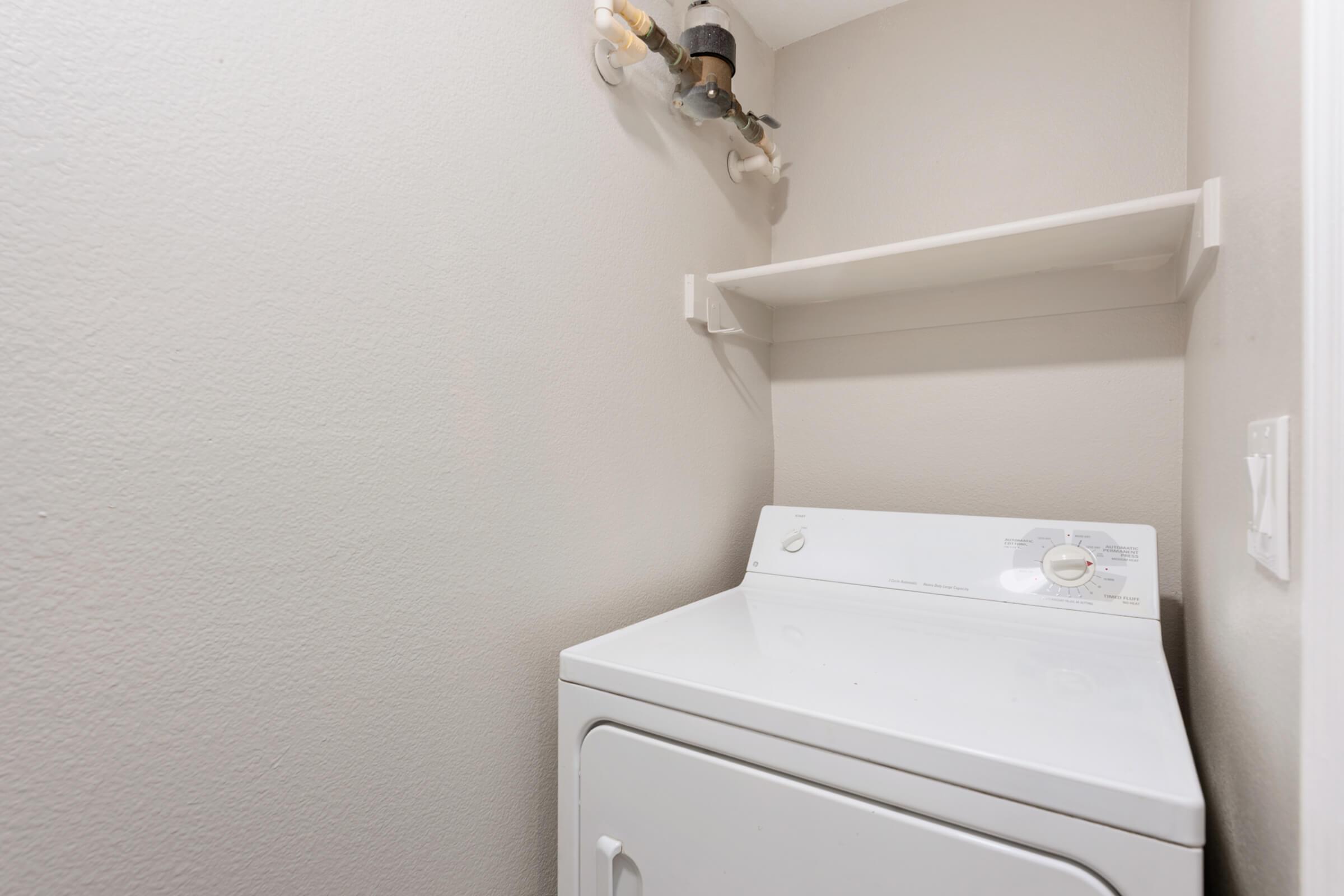 A small laundry room featuring a white washing machine against a neutral gray wall. Above the machine is a shelf for storage, and there are visible plumbing connections on the wall. The space is well-lit and tidy, emphasizing a minimalist design.