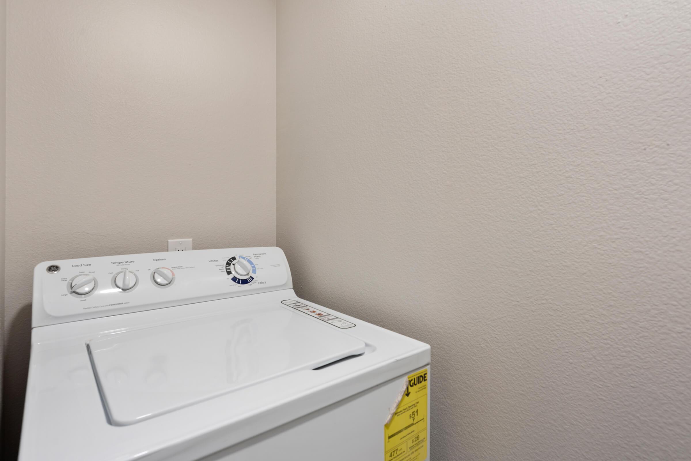 A white washing machine in a small laundry room with light gray walls. The machine features a control panel with dials and buttons. There is a safety label on the side of the washer. The space appears clean and uncluttered.