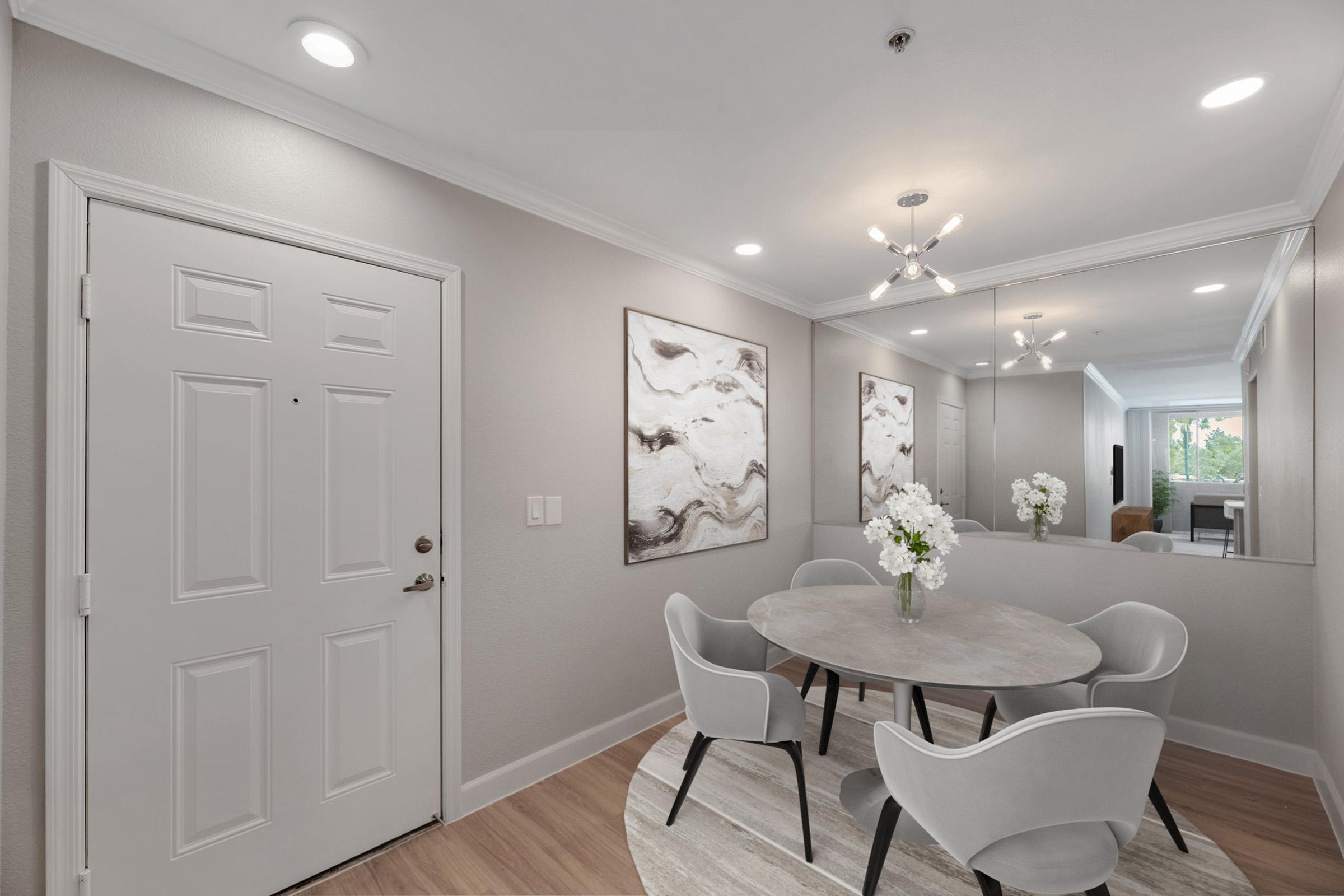 A modern foyer with a round dining table surrounded by four stylish chairs. The walls are painted in a light color, featuring two abstract art pieces. A mirror reflects the space, and a decorative light fixture hangs from the ceiling. Fresh white flowers are placed in the center of the table.