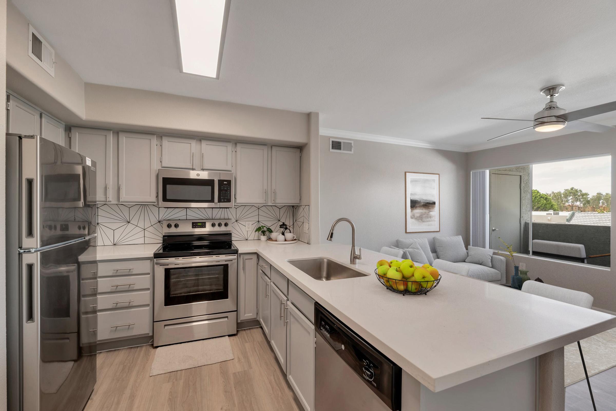 Modern kitchen with stainless steel appliances, including a refrigerator, oven, and microwave. Light gray cabinets and a white countertop. A small bowl of green apples on the island. Bright, open space with a living room visible in the background, featuring a couch and a ceiling fan. Natural light coming through a window.