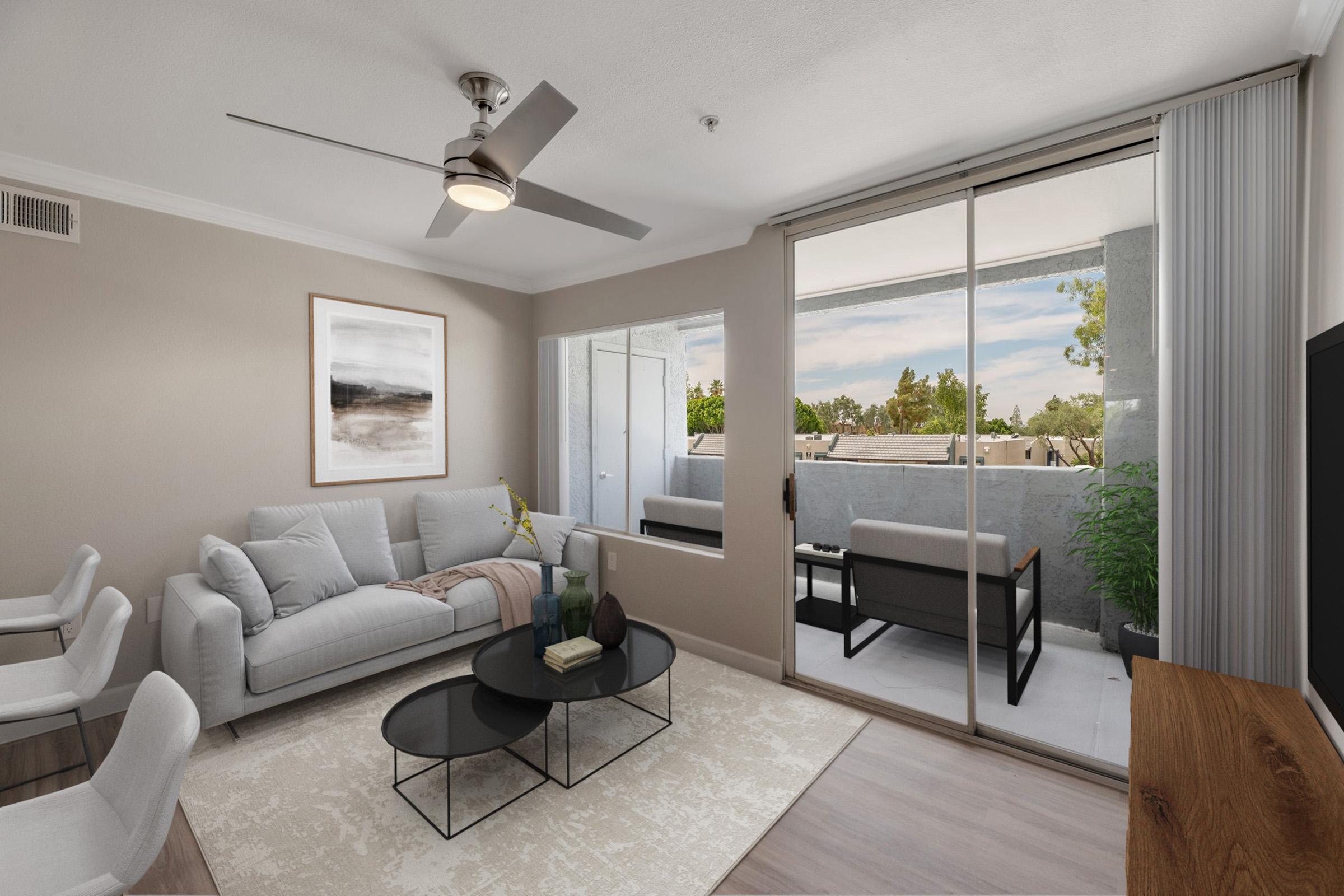 A modern living room featuring a light-colored sofa, a round black coffee table with decorative items, and a ceiling fan. Large windows allow natural light and offer a view of a balcony with outdoor seating. Neutral-toned walls and a minimalistic decor create a stylish, inviting atmosphere.