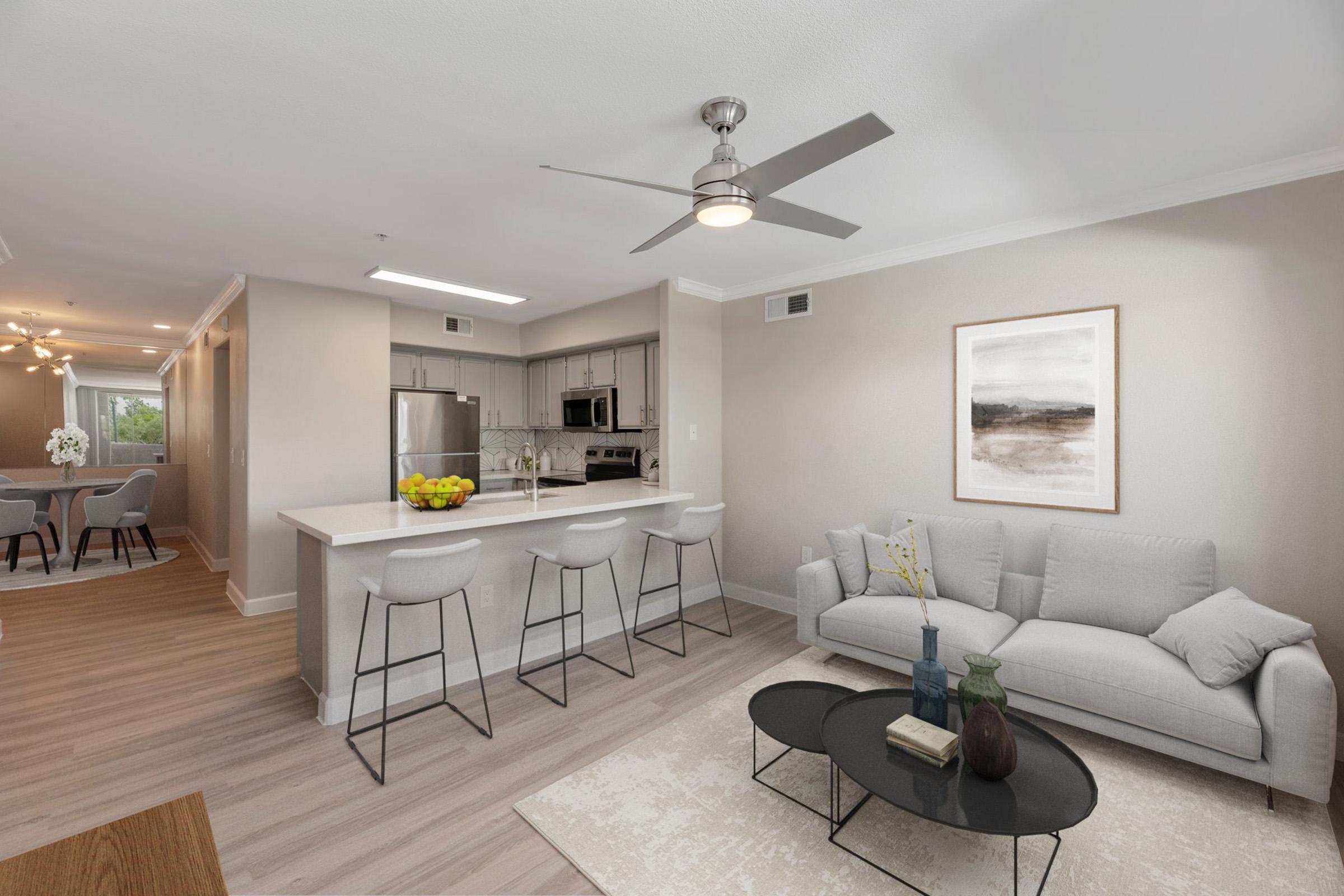 A modern living space featuring a light-colored sofa, a coffee table, and a ceiling fan. The open kitchen includes bar stools and stainless-steel appliances. Natural light fills the room, complemented by a simple artwork on the wall and a bowl of fruit on the counter. The flooring is a warm, light wood.