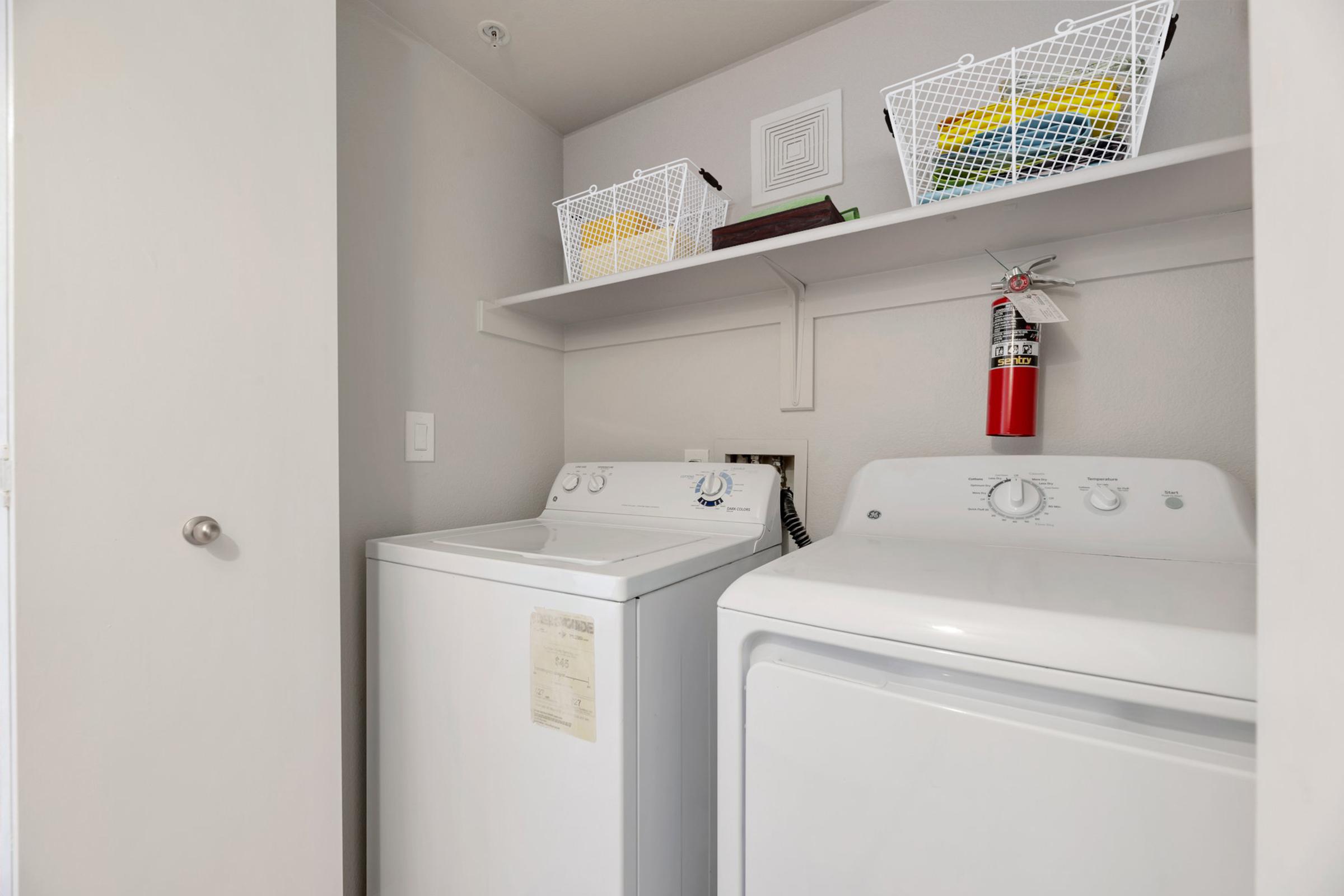 A laundry room featuring a white washing machine and dryer, both on an off-white floor. Above, there are three wire baskets on a shelf, containing various items. A fire extinguisher is mounted on the wall, providing safety equipment. The walls are painted a light color, contributing to a clean and organized appearance.