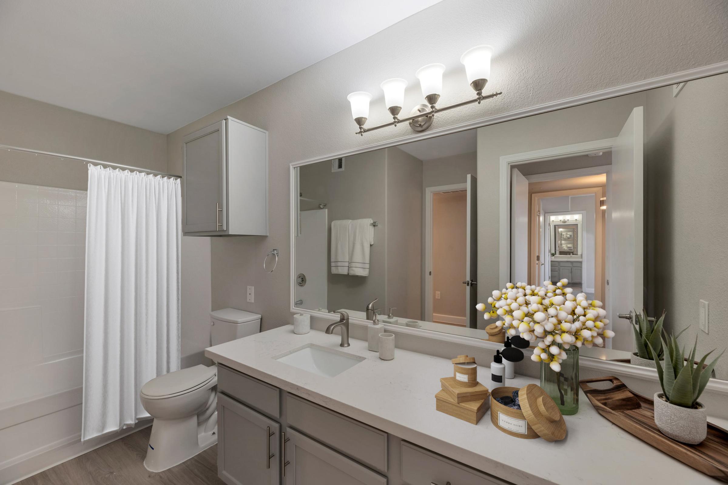 A modern bathroom featuring a white bathtub with a shower curtain, a long mirror with light fixtures above, gray cabinetry, a sink with a faucet, and decorative items including a flower arrangement and wooden trays. The room has neutral wall colors and leads to a hallway.