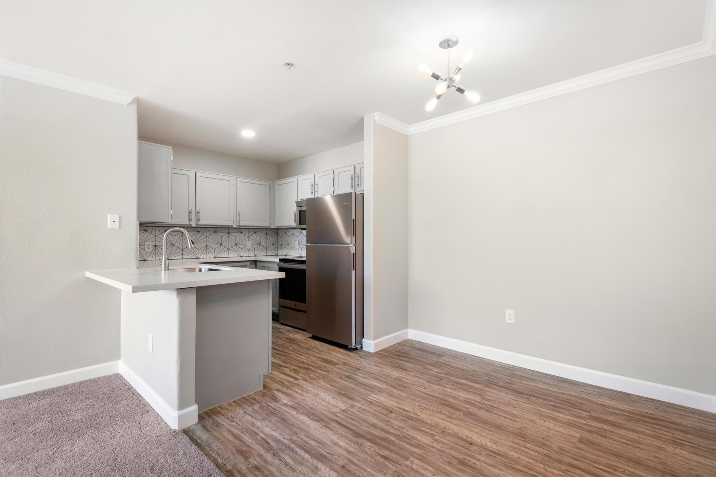 Modern kitchen with stainless steel appliances, including a refrigerator and oven. Light-colored cabinets are complemented by a patterned backsplash. The room features a pendant light fixture and wood-look flooring, with a carpeted area nearby. Neutral walls create a bright and spacious atmosphere.