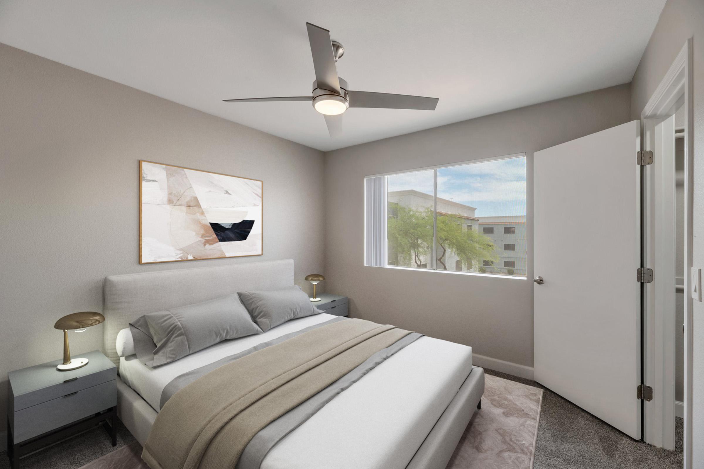 A modern bedroom featuring a gray upholstered bed with light gray bedding, two minimalist bedside tables with lamps, and a large window providing natural light. The walls are painted in soft gray tones, and there is a framed abstract artwork above the bed. A ceiling fan adds to the contemporary design.