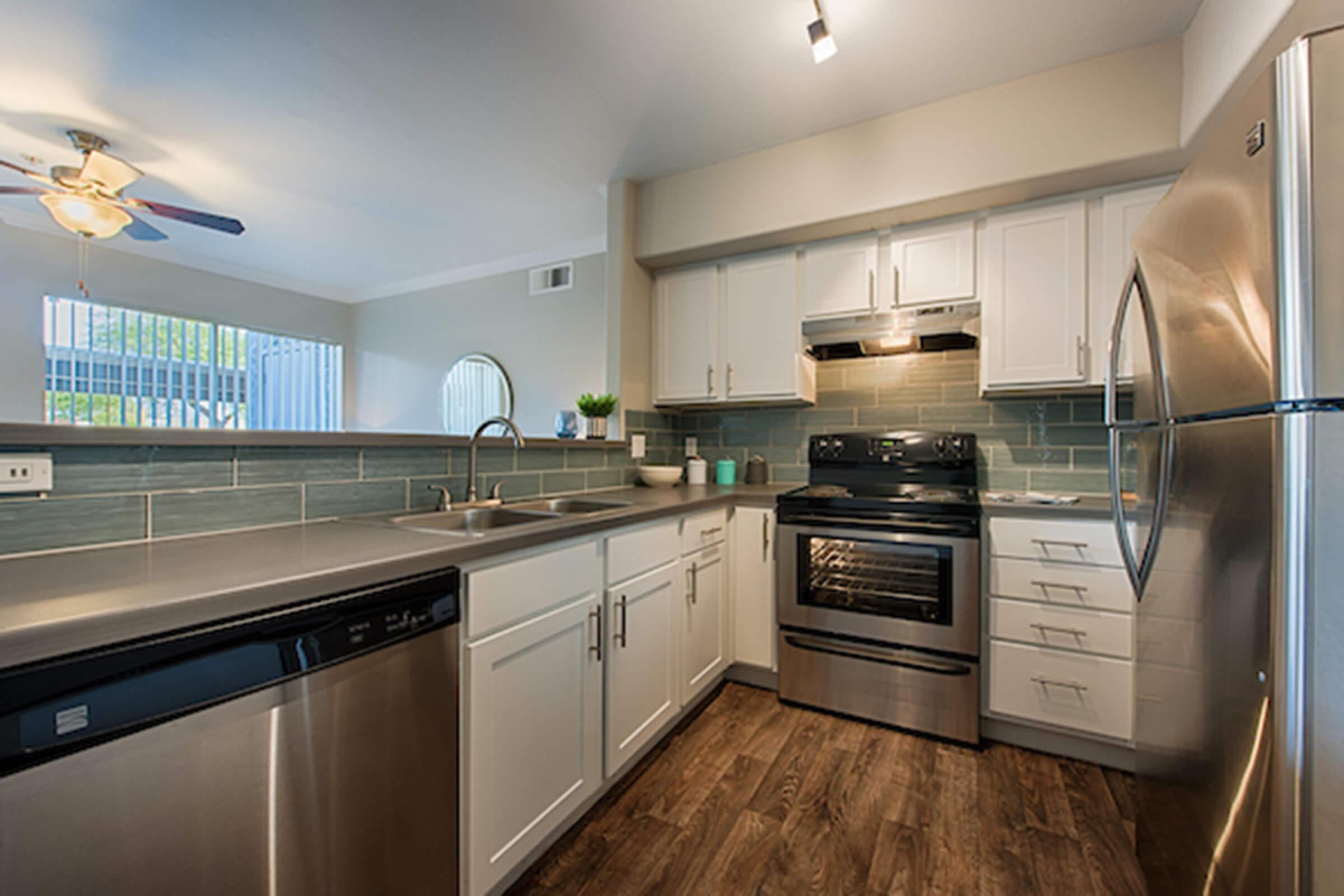 Modern kitchen featuring stainless steel appliances, including a dishwasher, oven, and refrigerator. The space has white cabinetry, a gray countertop, and a teal backsplash. Natural light comes from a window, creating a bright atmosphere, and there’s a ceiling fan for ventilation.