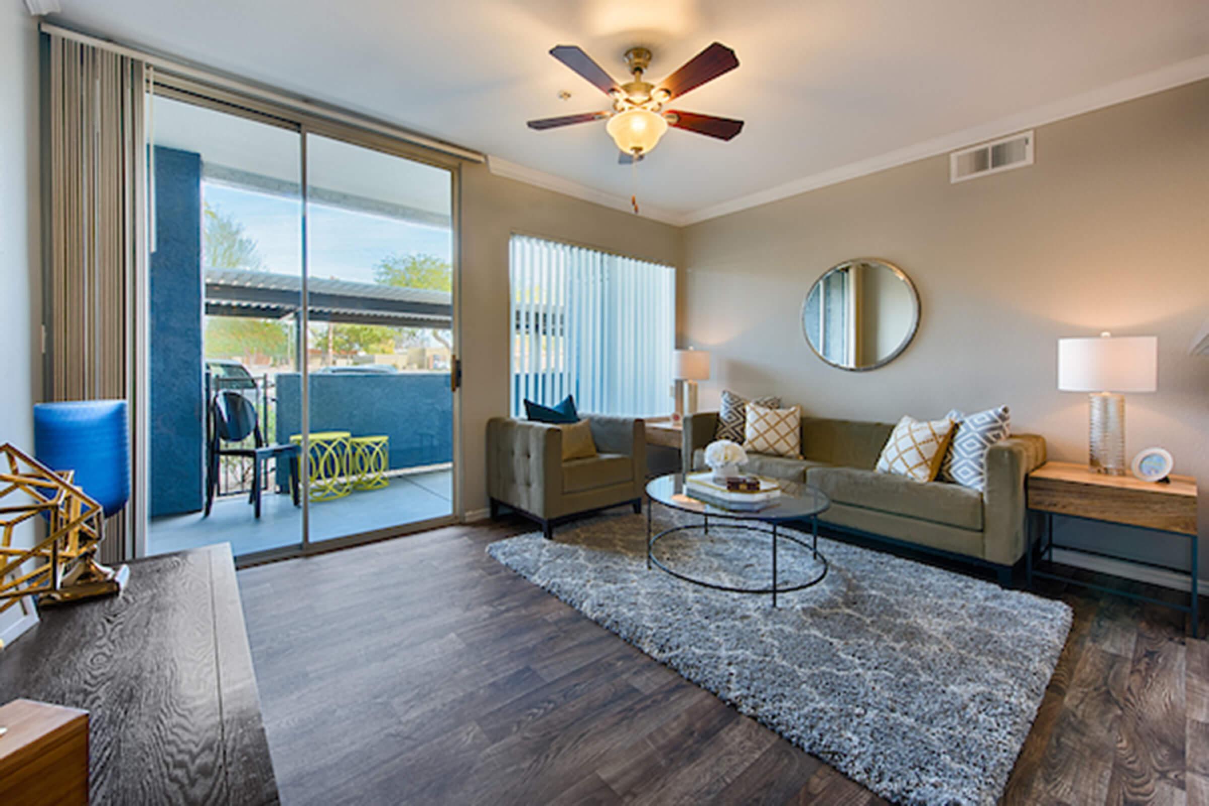 A modern living room featuring a comfortable sofa and armchair, a circular coffee table, and decorative pillows. A large window with sheer curtains lets in natural light, revealing an outdoor balcony. The floor is dark wood, and a stylish rug adds warmth to the space. Decorative accents enhance the contemporary aesthetic.