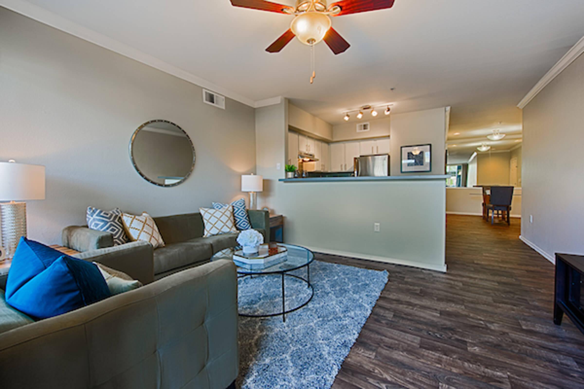 A cozy living room featuring a green sofa adorned with blue and patterned pillows, a round mirror on the wall, and a coffee table with decorative items. Natural light fills the space, highlighting the wooden flooring and a view into a well-lit kitchen and dining area in the background.