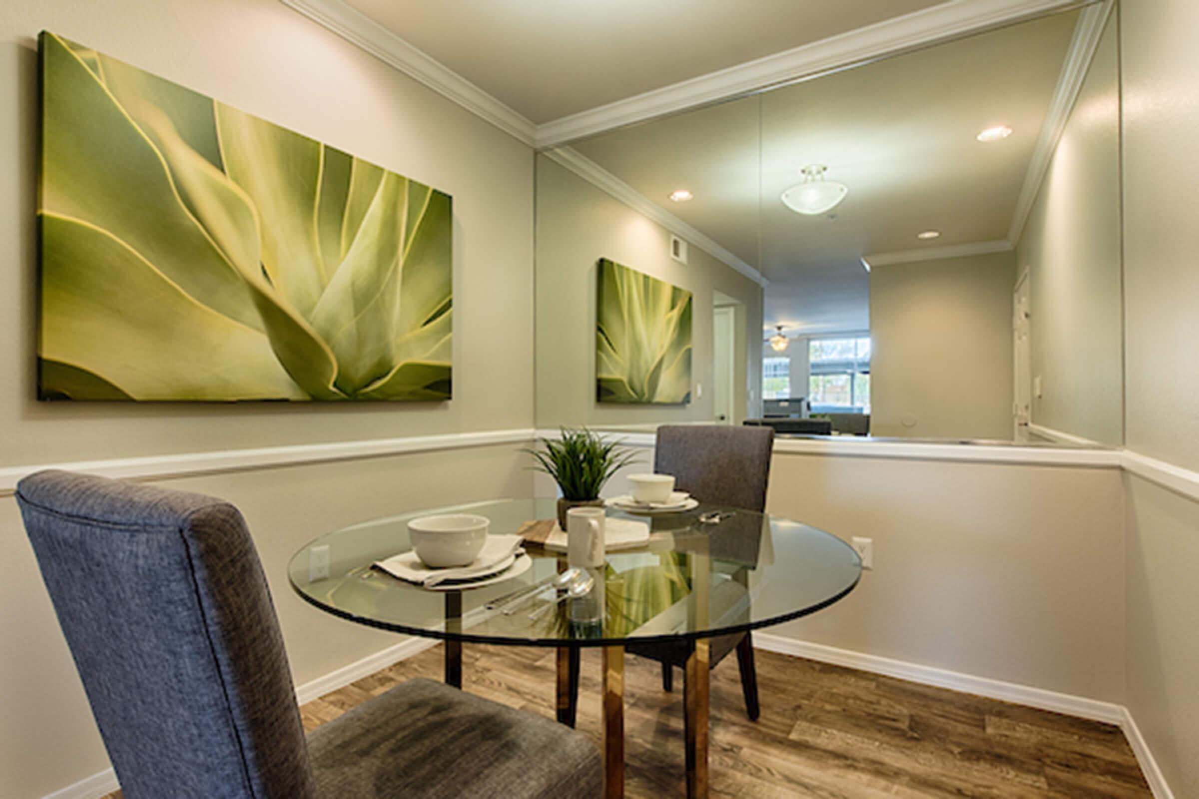 A cozy dining area featuring a round glass table set for two, with white bowls and a small vase. The walls are adorned with large green leaf artworks, and there's a mirrored wall reflecting the space. The floor is wooden, and the lighting is soft and inviting.