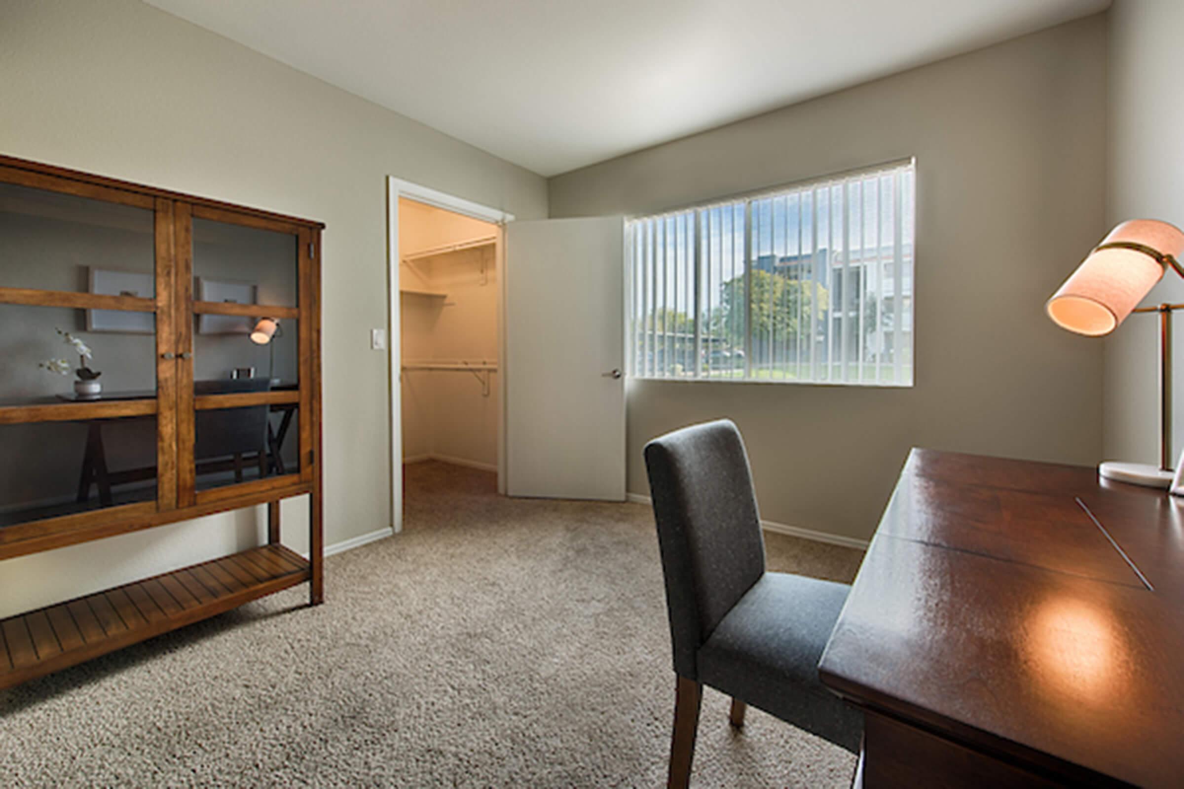 A well-lit room featuring a wooden desk and a gray chair. There is a glass-fronted cabinet against one wall, and a doorway leading to a spacious closet. The walls are painted a soft green, and a large window with vertical blinds allows natural light to fill the space. The carpet is beige.