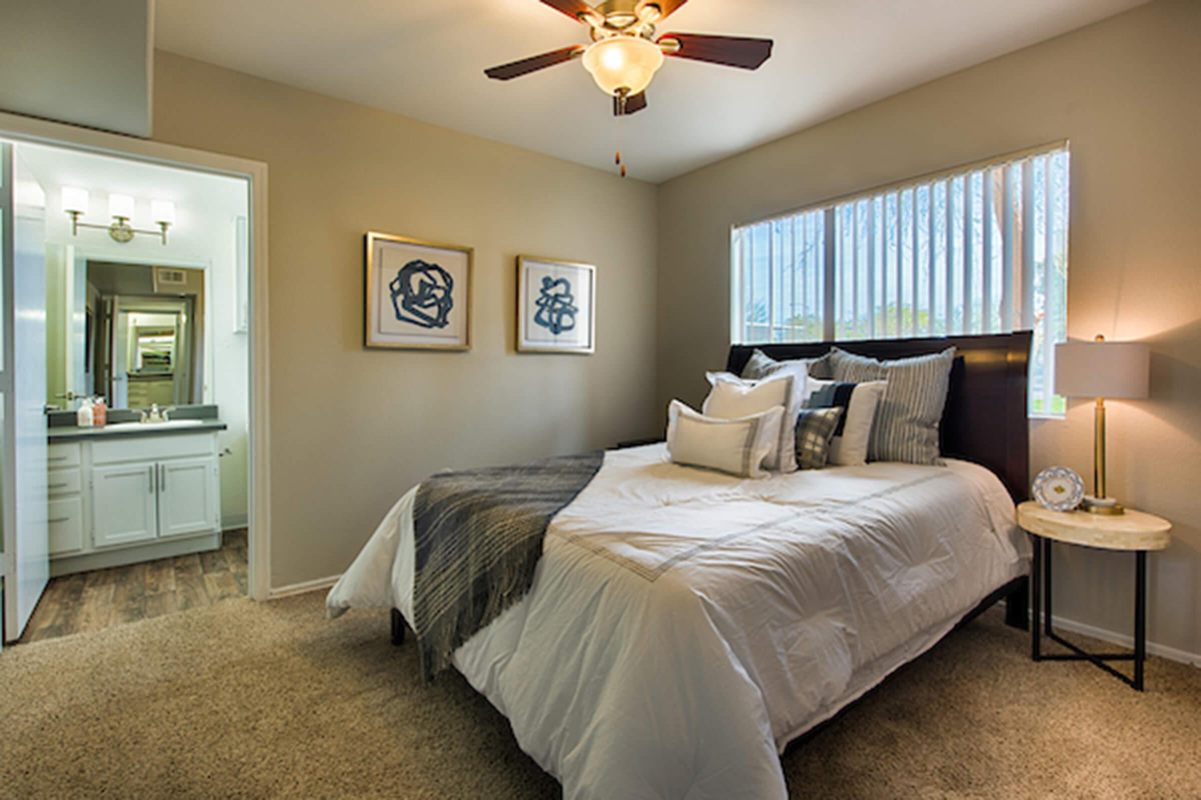 A cozy bedroom featuring a neatly made bed with decorative pillows, a ceiling fan, and two framed artworks on the wall. There's a bedside table with a lamp next to the bed, and a doorway leads to a modern bathroom with white cabinetry visible in the background. Natural light filters through a window with blinds.