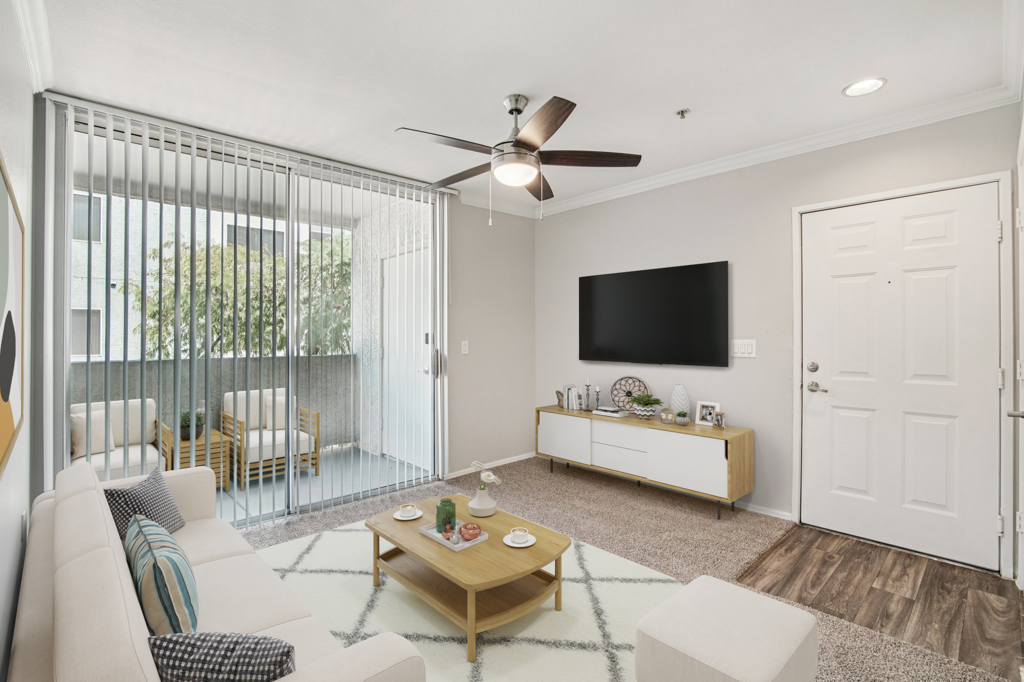 A modern living room featuring a light-colored sofa with decorative pillows, a wooden coffee table with snacks, a flat-screen TV mounted on a wall, and a door leading to an outdoor balcony. Floor-to-ceiling sliding glass doors provide natural light, and there are cozy seating options visible on the balcony.