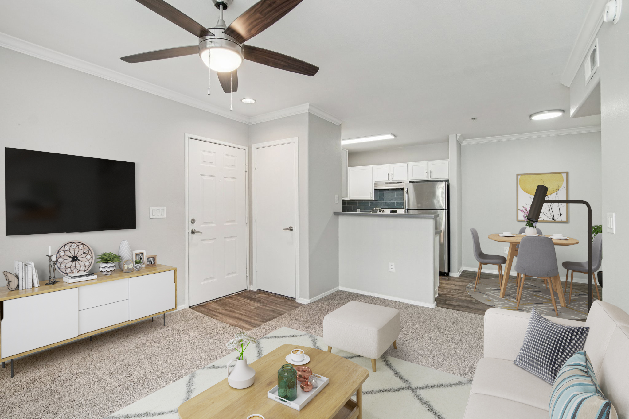A modern living room featuring a light-colored couch, a wooden coffee table, and a flat-screen TV mounted on the wall. The space is accented with a ceiling fan and includes a dining area with a round table and chairs. The decor is minimalistic and inviting, with neutral tones and a cozy rug.