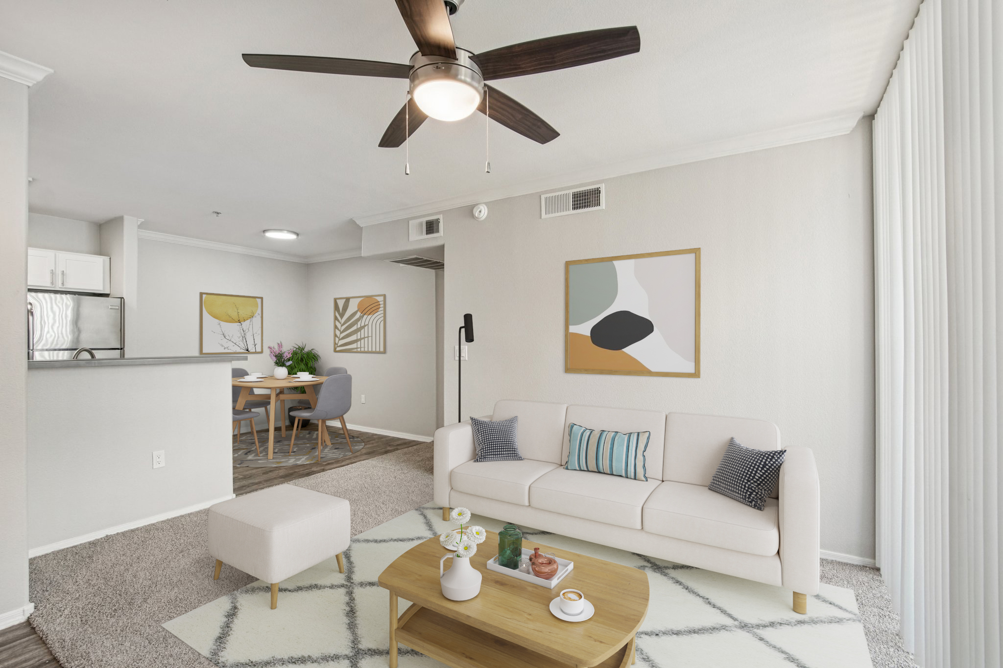 A modern living space featuring a light-colored sofa with decorative pillows, a round coffee table with snacks, and a ceiling fan. Artwork adorns the walls, and there's a dining area visible in the background. The floor is carpeted, enhancing the cozy ambiance of the room.