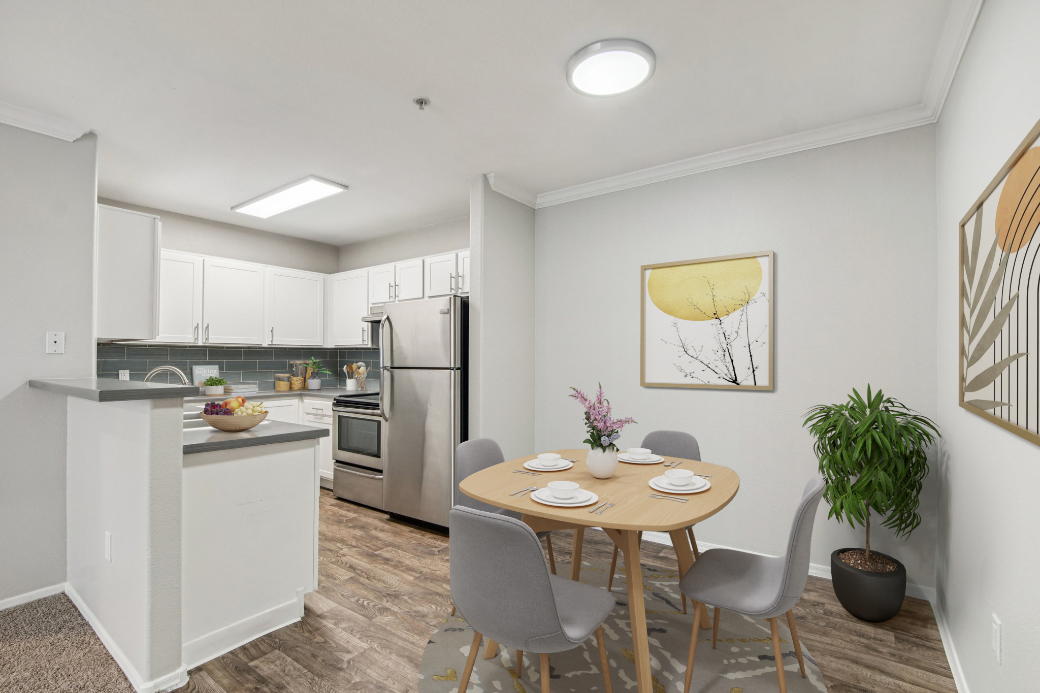 A modern kitchen and dining area featuring a round wooden table set for four, with gray chairs. The kitchen includes white cabinets, stainless steel appliances, and a gray backsplash. A small plant and vase with flowers are on the table, and a wall art piece with a yellow circle adds a decorative touch.