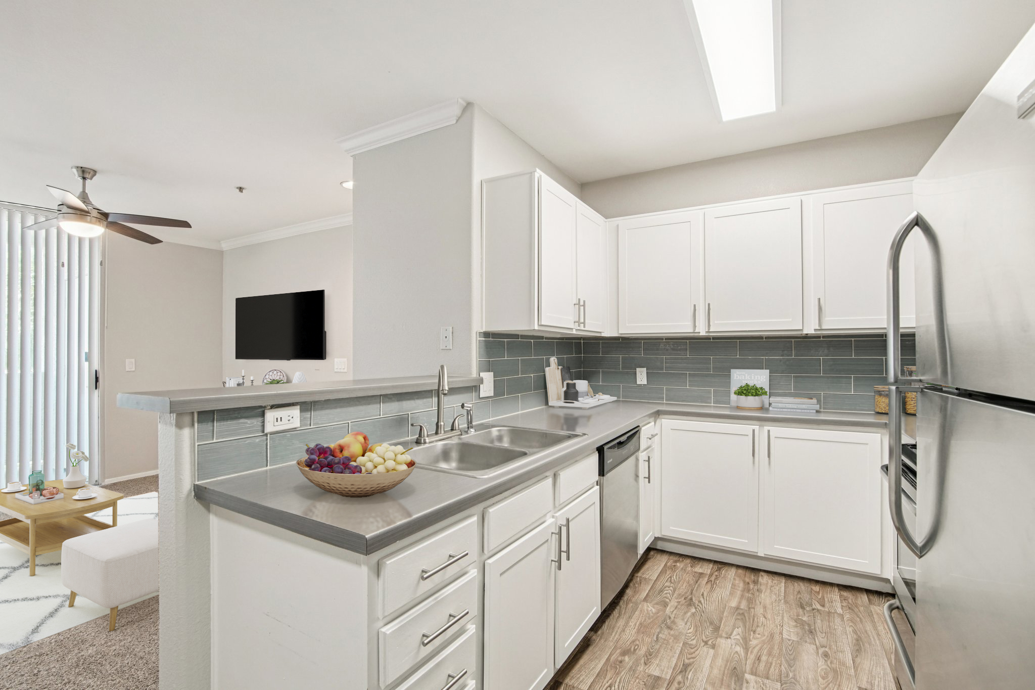 Modern kitchen featuring white cabinets, stainless steel appliances, and a gray tile backsplash. A sink with a fruit bowl sits on the counter. The space is well-lit with a ceiling fan and a living area visible in the background, displaying a television and cozy seating. Wood-like flooring adds a warm touch.