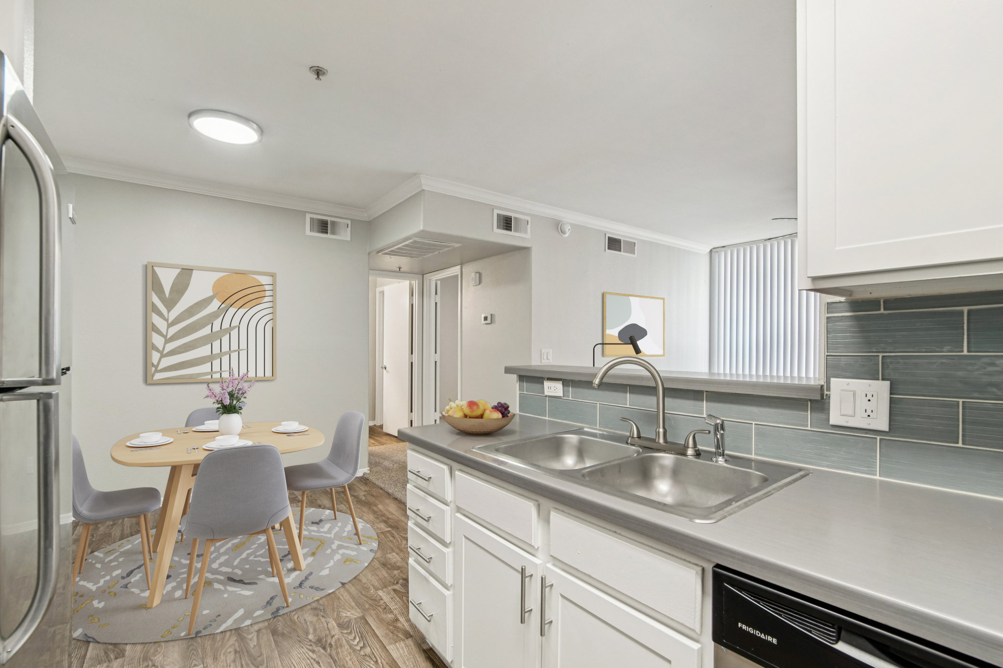 A modern kitchen featuring stainless steel appliances, a double sink, and light-colored cabinetry. In the background, a dining area with a round table and four chairs is visible, surrounded by natural light. Decorative elements include a vase with flowers and fruit on the table, enhancing the inviting atmosphere.