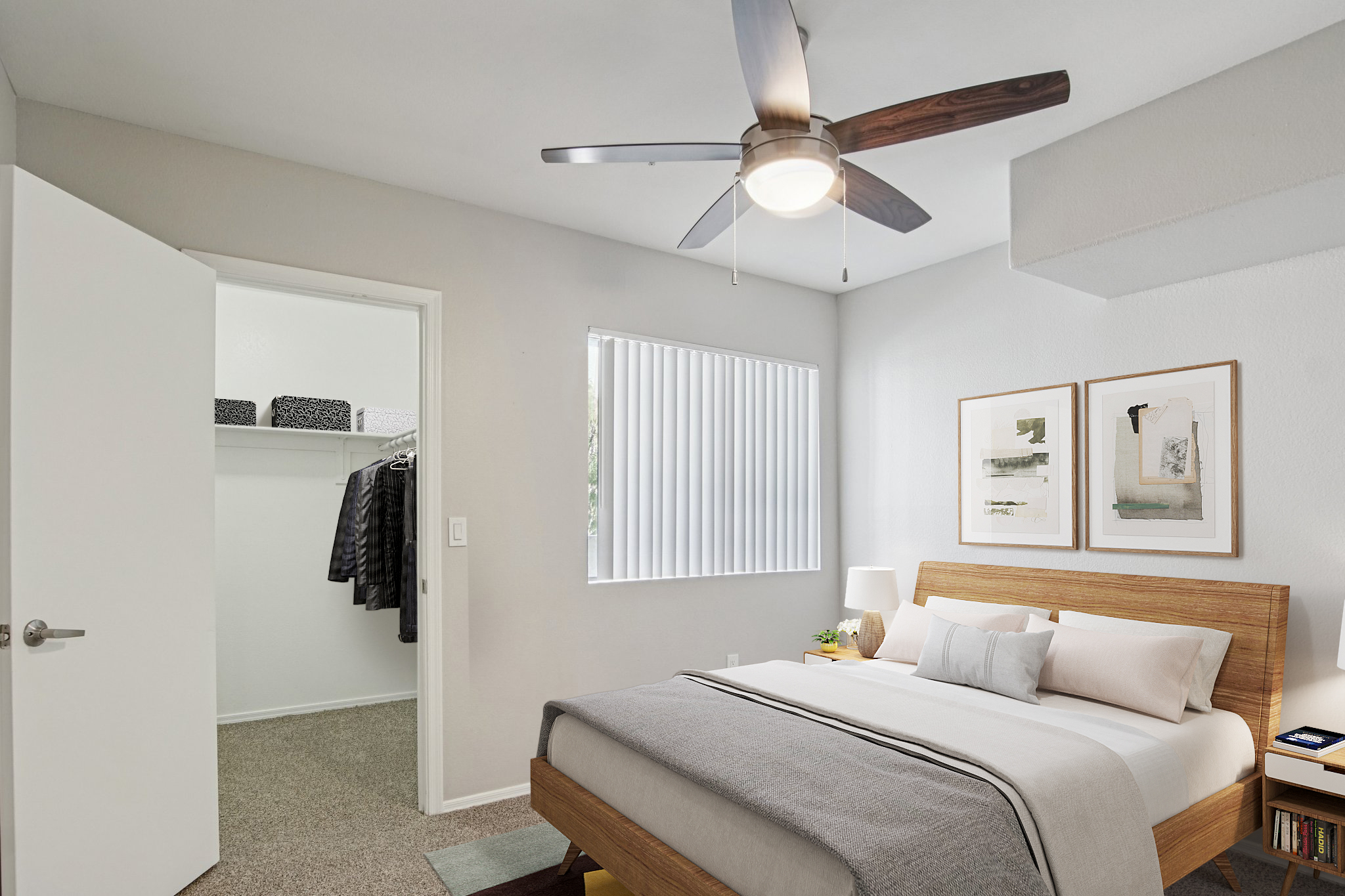 A modern bedroom featuring a bed with a light wood frame and gray bedding, a ceiling fan, and two framed artworks on the wall. There is a window with blinds providing natural light, and a closet with neatly arranged storage boxes. The room has neutral gray walls and a cozy carpeted floor.