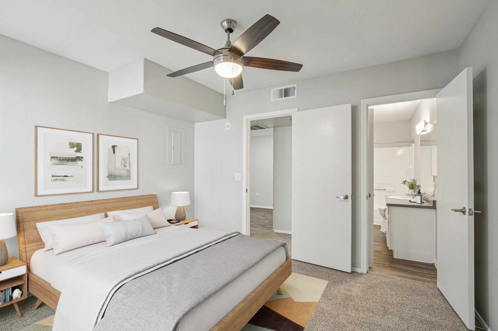 A modern bedroom featuring a large bed with neutral bedding, two bedside lamps, and framed artwork on the walls. A ceiling fan is installed above, and a door leads to a bathroom area. The room has light gray walls and a stylish, colorful rug on the floor, with a spacious closet visible.