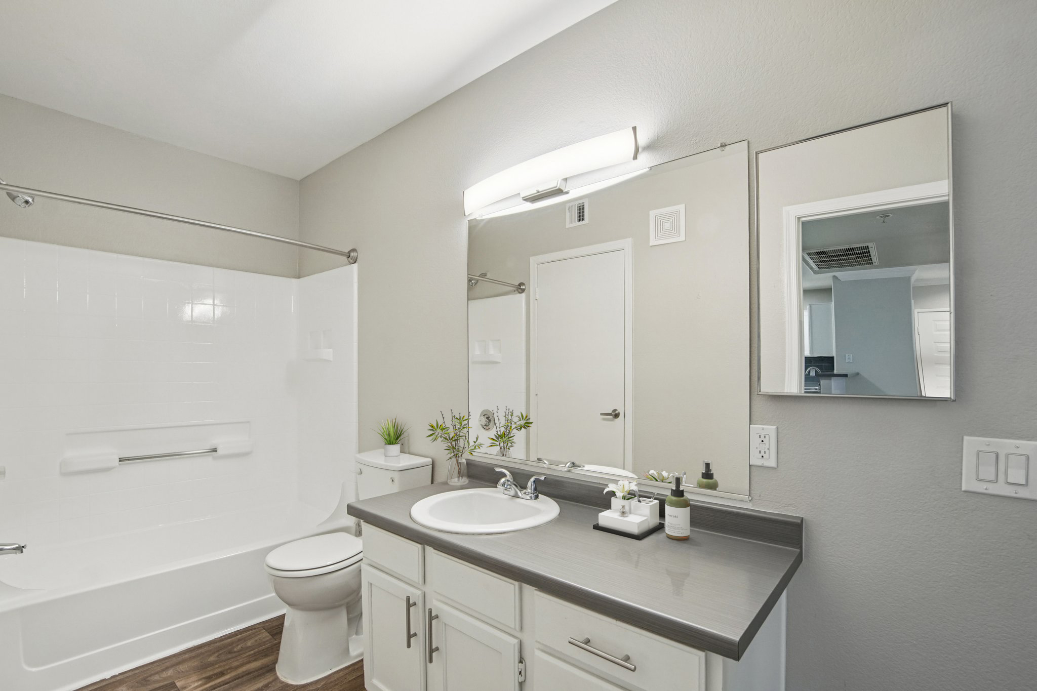 Modern bathroom featuring a white bathtub with a shower, a sleek countertop with a sink, a mirror, and minimal decorative elements. Neutral wall colors and wooden flooring create a clean and inviting atmosphere. Natural light enhances the space, making it bright and open.